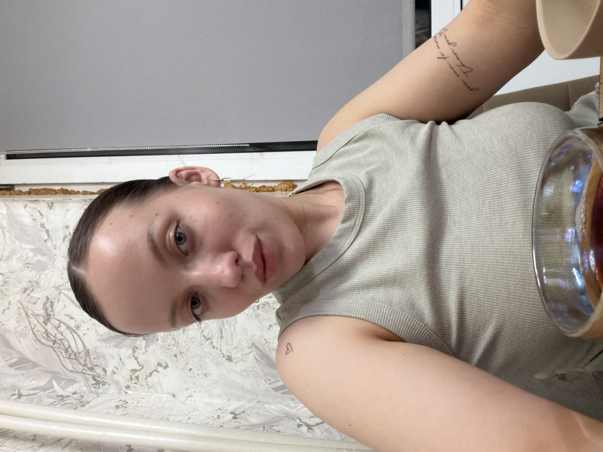 The image is a sideways, close-up selfie of a young woman taken indoors, with her head oriented towards the left and her body extending to the right. She has dark hair pulled back, light-colored eyes (appearing blue or grey), and a neutral expression as she looks directly at the camera.
She is wearing a light green or grey ribbed tank top. On her left shoulder/upper arm, there is a small, outlined heart tattoo. On her right forearm, a script tattoo is visible, which appears to read "all of me loves all of you". A delicate gold chain is also visible around her neck.
To her right, a glass containing a dark beverage, possibly tea or coffee, is partially visible. The background features a white wallpaper with intricate, light grey or metallic leaf-like patterns. Behind her head and shoulder, a window with a light grey roller blind is visible. The window sill underneath the blind contains some dried, brownish, textured debris or material.
The scene depicts a casual indoor setting, likely a home, during daytime. The location context provided is Reshetnikovo, Russia.