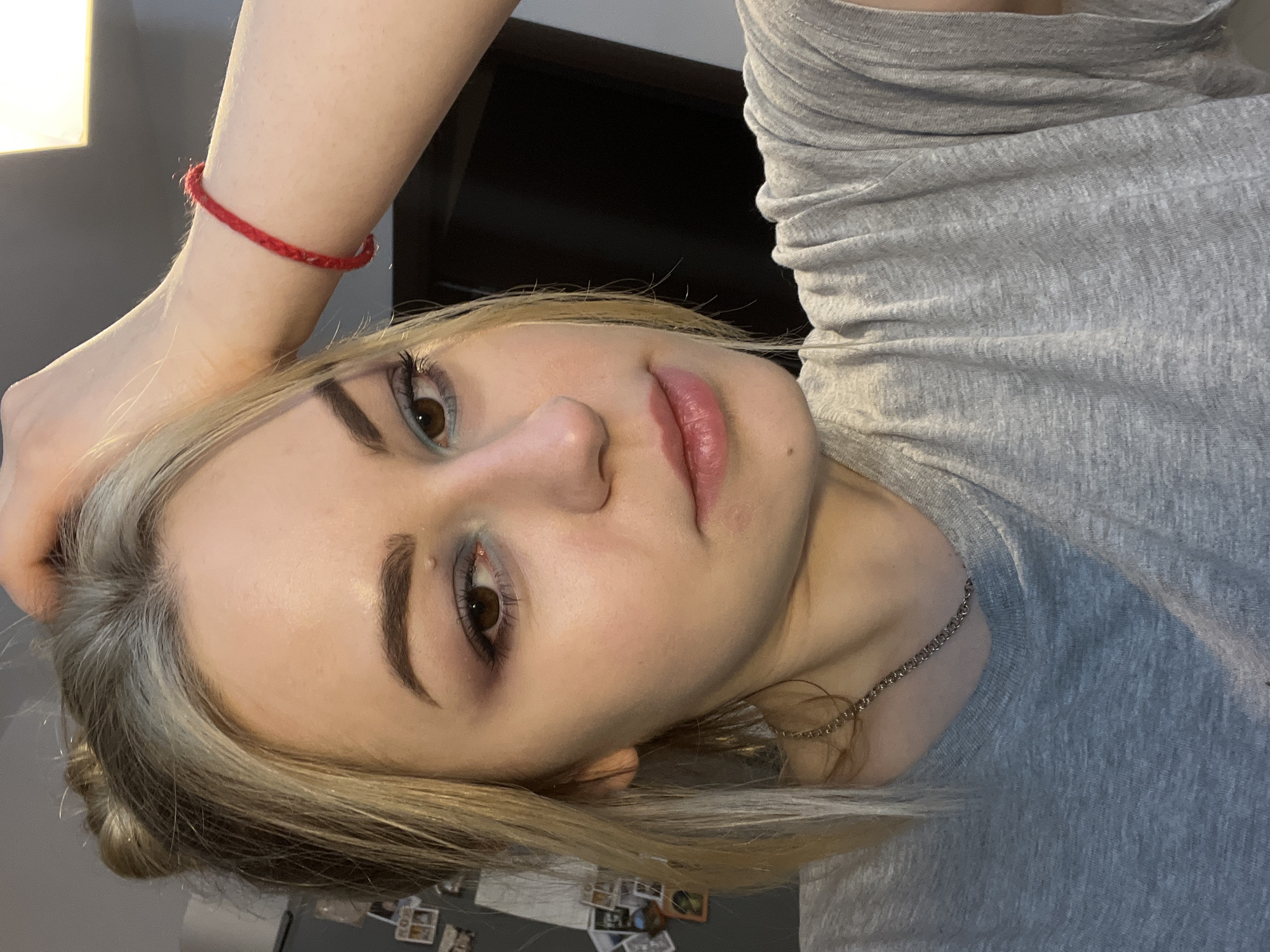 A fair-skinned individual with blonde and gray hair is shown in a close-up, selfie-style shot. The person is wearing a light gray t-shirt and a thin silver chain around their neck. On their wrist is a red string bracelet. They have light blue eyeshadow and a neutral expression. The background appears to be an interior setting with a light source above and a dark object behind the person's head. There is a refrigerator door visible in the bottom of the frame with several magnets and pictures affixed. This image does not contain visual clues to specify its location as Boreti, Montenegro.