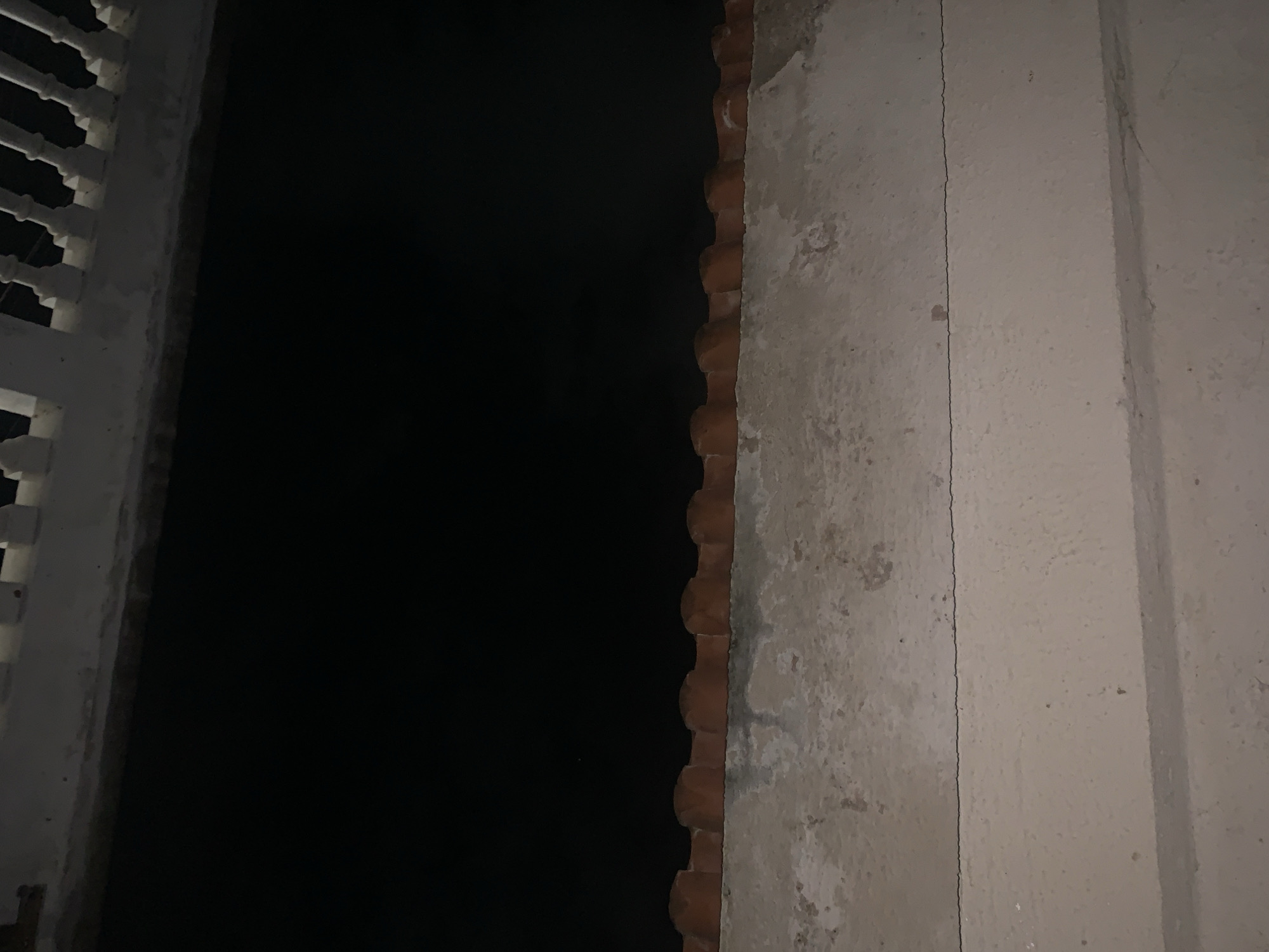 This media file presents a dark, static scene, likely captured at night from within a structure in Santa Ana, Venezuela. On the far left, a portion of a white balustrade or decorative railing is visible, featuring several distinct, ornate vertical pillars against the darkness. The central and upper-left part of the image is dominated by a vast expanse of deep black, indicating either profound darkness or an unlit open space beyond the structure.
Moving towards the middle-right, a vertical band of reddish-brown, textured material is prominent, resembling the curved edges of traditional clay roof tiles or bricks arranged vertically. This forms a rough boundary with the dark background. To the right of this, a light-colored, textured wall, likely made of concrete or stucco, fills the remainder of the frame. This wall shows signs of age or wear, with some discoloration and a noticeable, thin vertical crack running down its surface. Further to the right, a slightly darker vertical section suggests a structural corner or an adjacent wall. No people are visible, and no specific activity or event is taking place. The overall impression is one of architectural detail in very low light conditions, highlighting textures and structural elements. No text is visible in the image.