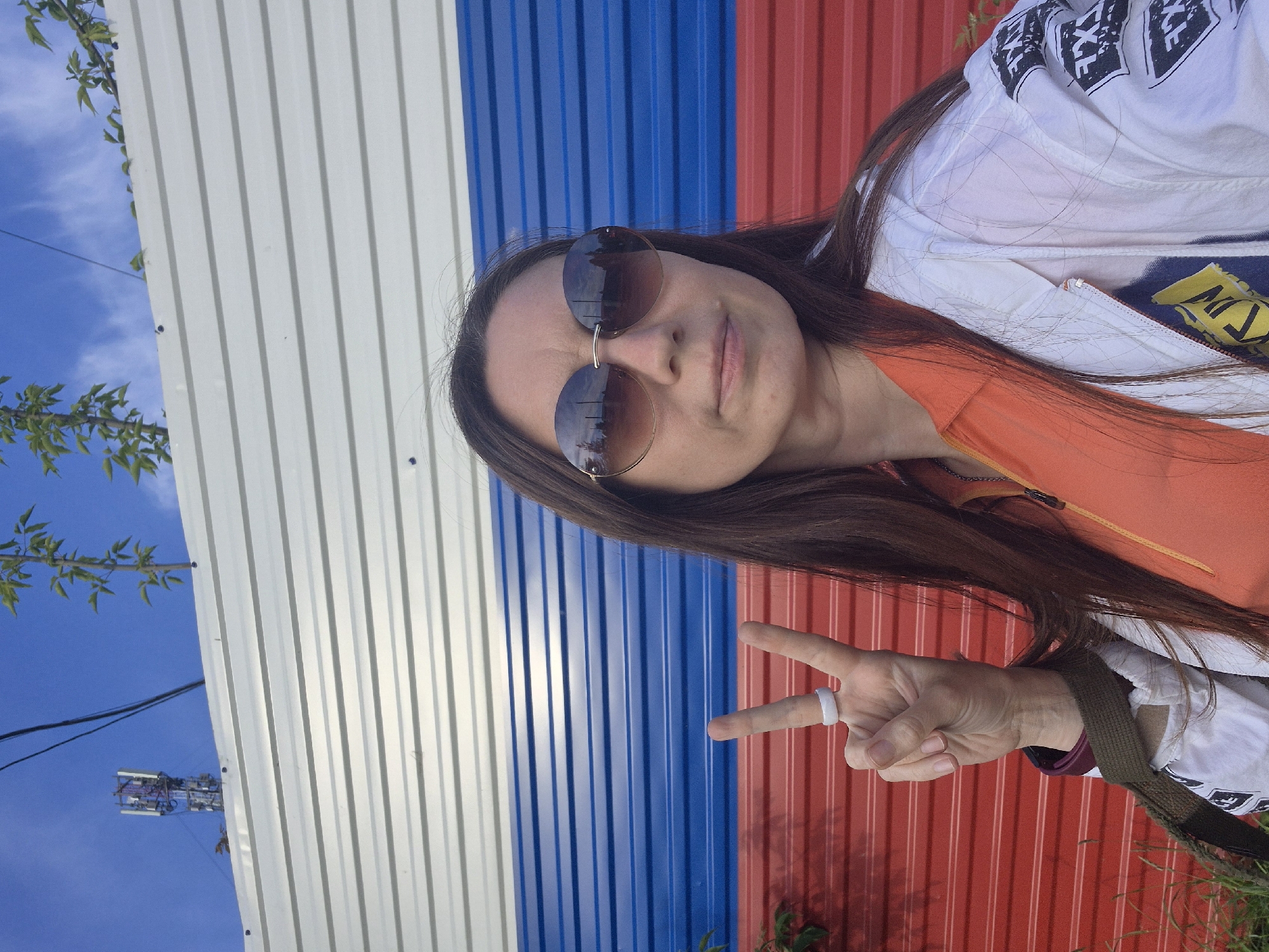 This media file captures a selfie-style portrait of a young woman posing outdoors, set against a backdrop featuring the colors of the Russian flag. The image is oriented vertically but appears to have been taken horizontally and then rotated 90 degrees clockwise.
The subject is a woman with long, dark brown hair, wearing large, round sunglasses that reflect the sky and surroundings. She has a neutral to slightly smiling expression. She is dressed in an orange top underneath a white jacket that has black and white graphic patterns on the shoulders and sleeves, including some illegible text-like designs. A dark olive green strap, possibly from a bag, is visible across her right shoulder. Her right hand is raised, forming a peace sign, and she wears a white ring on her right ring finger.
The backdrop consists of a corrugated metal wall painted with distinct vertical stripes of white, blue, and red, strongly resembling the national flag of Russia, which aligns with the provided location context of Chaikovsky, Russia. To the left of the wall, a bright, clear blue sky is visible, with a few wispy clouds and green foliage from a tree. In the distance, a communication tower or cellular mast stands out against the sky, along with thin black power lines.
The bright, even lighting indicates it is daytime with clear weather. The overall mood conveyed by the woman's pose and expression, coupled with the peace sign, suggests a relaxed and positive atmosphere.