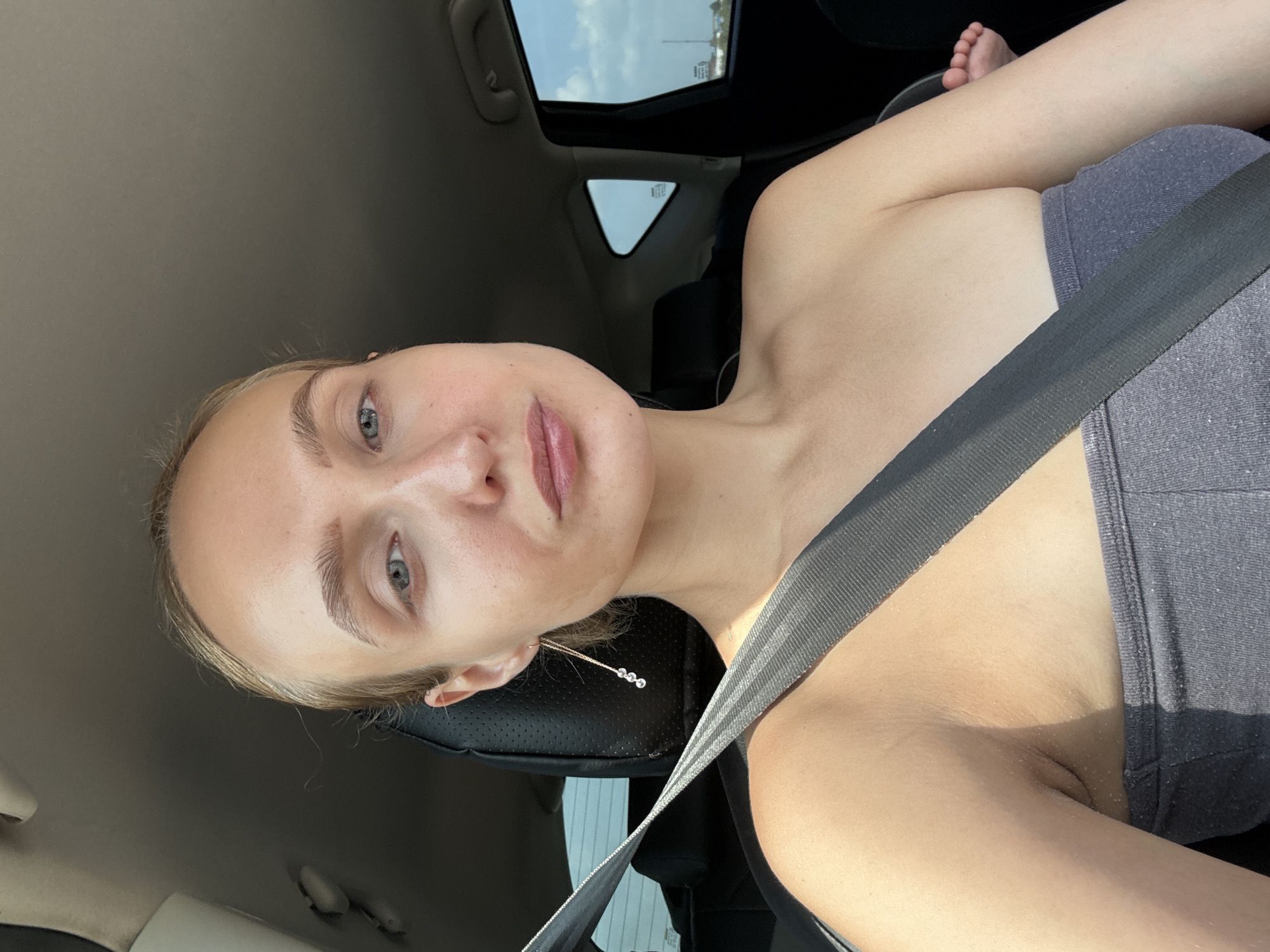 In a car, a fair-skinned woman with blonde hair pulled back is seated in the driver's seat. She is looking directly at the camera, her face tilted slightly upwards. She is wearing a grey strapless top and a prominent black seatbelt crosses her chest. To her right, a baby's foot is visible, suggesting the baby is in a car seat. The background shows the interior of the car, including a headrest and a window with a view of a cloudy sky and distant buildings. The woman is wearing a long, dangling earring with three small beads. The image appears to have been taken during the daytime, with natural light illuminating the scene. The context of Pirogovo, Russia is not derivable from the visual information provided in the image.