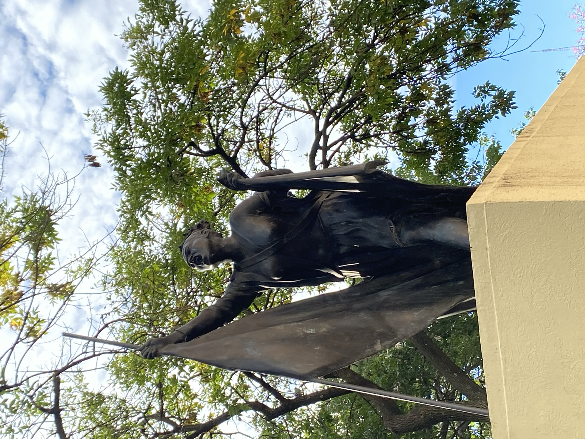 The image presents a low-angle view of a bronze statue mounted on a stone structure, situated in an outdoor environment with trees overhead. The statue depicts a figure in what appears to be battle clothing, holding a flag or banner. The weathered texture of the bronze suggests the statue is aged. The stone structure is a light tan color. The background features green foliage from the trees, which partially obscures a blue sky with scattered white clouds.
While the location is said to be Buenos Aires, Argentina, no element in the image allows direct determination of its specific city location. Without further information, the specific location in Buenos Aires cannot be confirmed from the visual elements of the image alone.