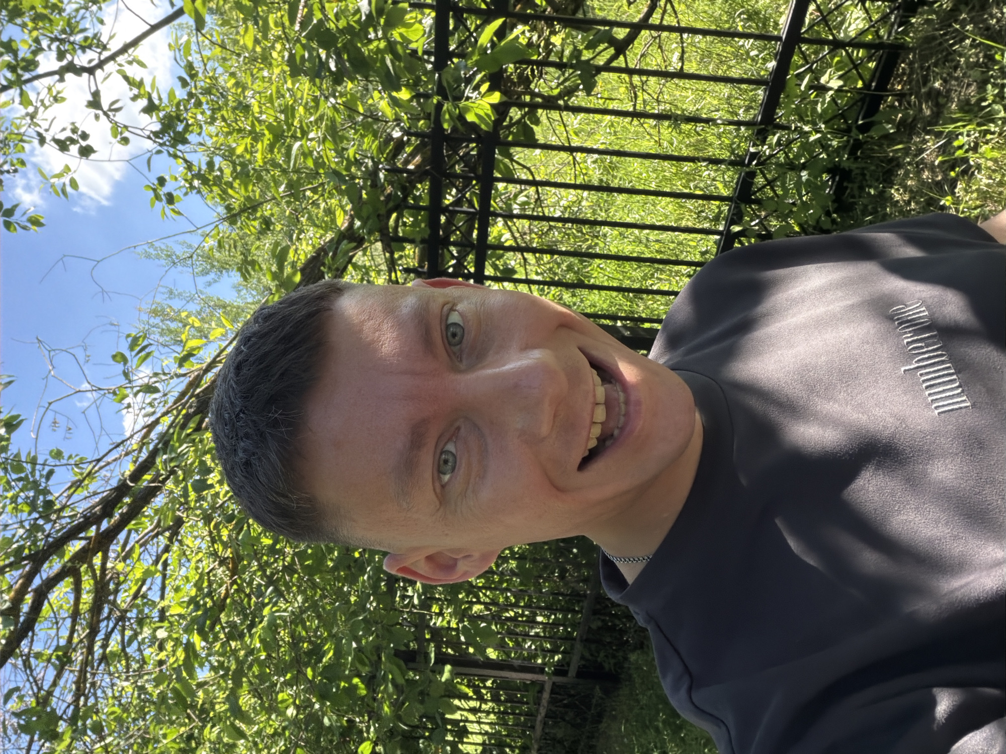 An outdoor, close-up, eye-level selfie shows a smiling person from the chest up, positioned in the center of the frame. The person, who appears to be male, has short, dark hair, light skin, and blue eyes. He is wearing a dark grey t-shirt with a silver chain necklace. His mouth is open, revealing teeth, and he is looking directly at the camera.
Behind the person, a black metal fence runs horizontally across the mid-ground. Beyond the fence, lush green foliage and trees dominate the background, with a bright blue sky with scattered white clouds visible above. The lighting suggests it is daytime, likely sunny. The setting appears to be a park or a garden.