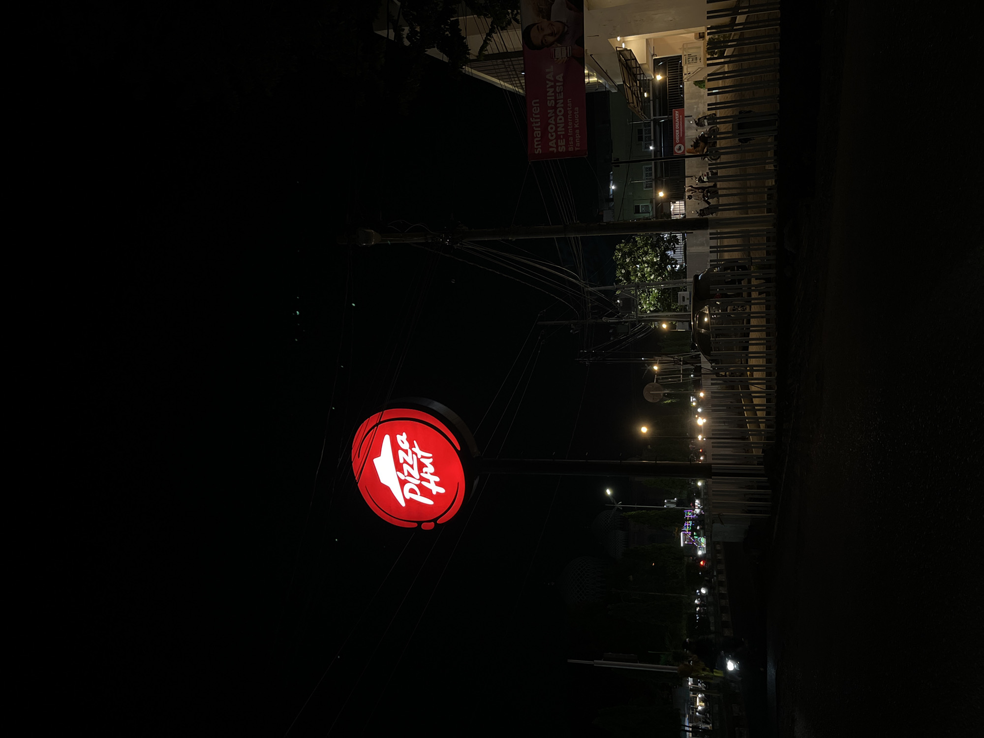 A red, circular Pizza Hut sign is illuminated against a dark night sky. The sign features the white Pizza Hut logo, which includes a stylized red roof and the text "Pizza Hut." The sign is mounted on a tall, dark pole. To the right of the sign, a building is visible with a banner that reads "smartfren" and "JAGOAN SINYAL SE-INDONESIA BISA INTERNETAN TANPA KUOTA." Several streetlights are also visible, casting a warm glow on the surroundings. In the foreground, a road is visible with a few vehicles, including a motorcycle with its headlights on. A metal fence runs along the edge of the property. The scene appears to be on a street in the city of Lhokseumawe, Indonesia, likely at night.