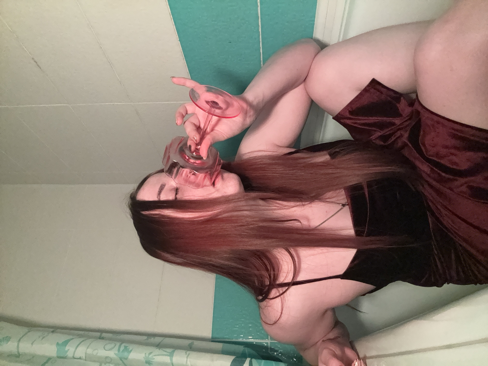 A woman is reclining in a bathtub, drinking from a glass. She has long red hair and is wearing a dark velvet dress. The bathtub is in a bathroom with tiled walls. The location is Izhevsk, Russia.