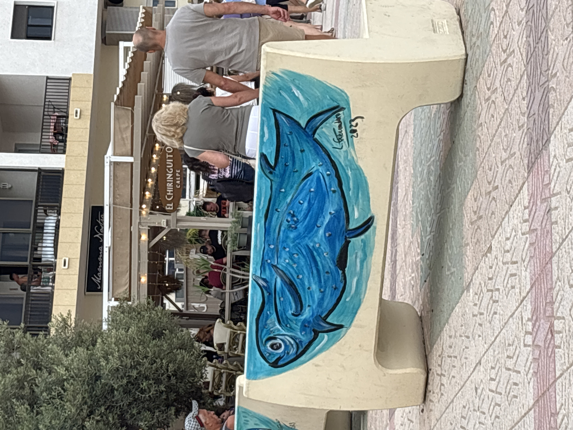 The image depicts a concrete bench decorated with a painting of a blue fish. The bench is positioned outdoors on a patterned pavement. In the background, a building with multiple levels and balconies is visible. Several people are present, with some walking in the background and others seated at tables in an outdoor seating area. A sign for "El Chiringuito Calpe" is also visible in the background. The overall setting appears to be a public space, possibly a promenade or a street in a town near the sea. The lighting suggests it is daytime.