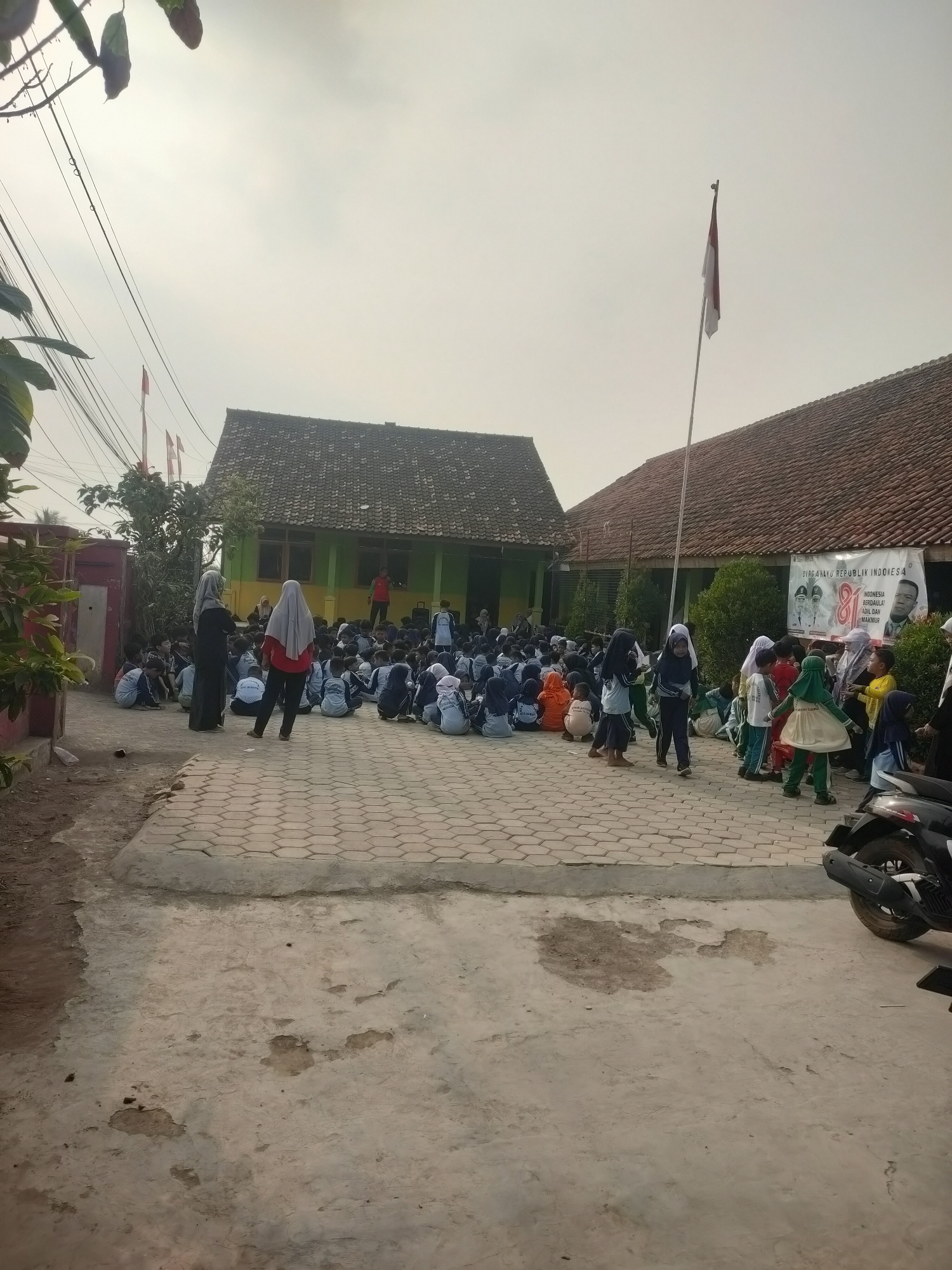 The image depicts an outdoor scene at what appears to be a school in Subang, Indonesia. A large group of children, many wearing school uniforms and hijabs, are seated on the ground in an open paved area. Several adults, also in modest attire, are standing among them or observing. Behind the children are two school buildings with tiled roofs. A large banner is visible on the right-hand building, featuring Indonesian flags and portraits, possibly celebrating a national holiday. A flagpole with a red and white Indonesian flag stands in front of the building on the right. The environment suggests a sunny day, with a clear sky and some shadows cast on the ground. The foreground shows a rough concrete surface and a parked motorcycle. The overall atmosphere suggests a community or school gathering.