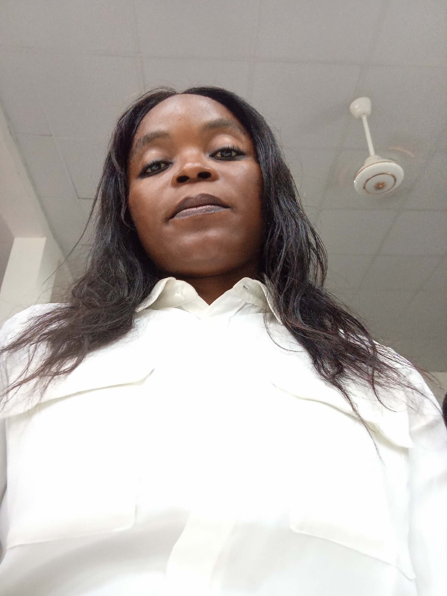 A low-angle, medium shot captures a Black woman from the chest up, looking directly into the camera. She has dark hair that falls past her shoulders and is wearing a white collared shirt. Her makeup includes dark eyeliner and lipstick. The background is a ceiling with a white ceiling fan. The image appears to be taken indoors in a brightly lit room. The location in the city of Kason, Nigeria, is not discernible from the visual information provided in the image.