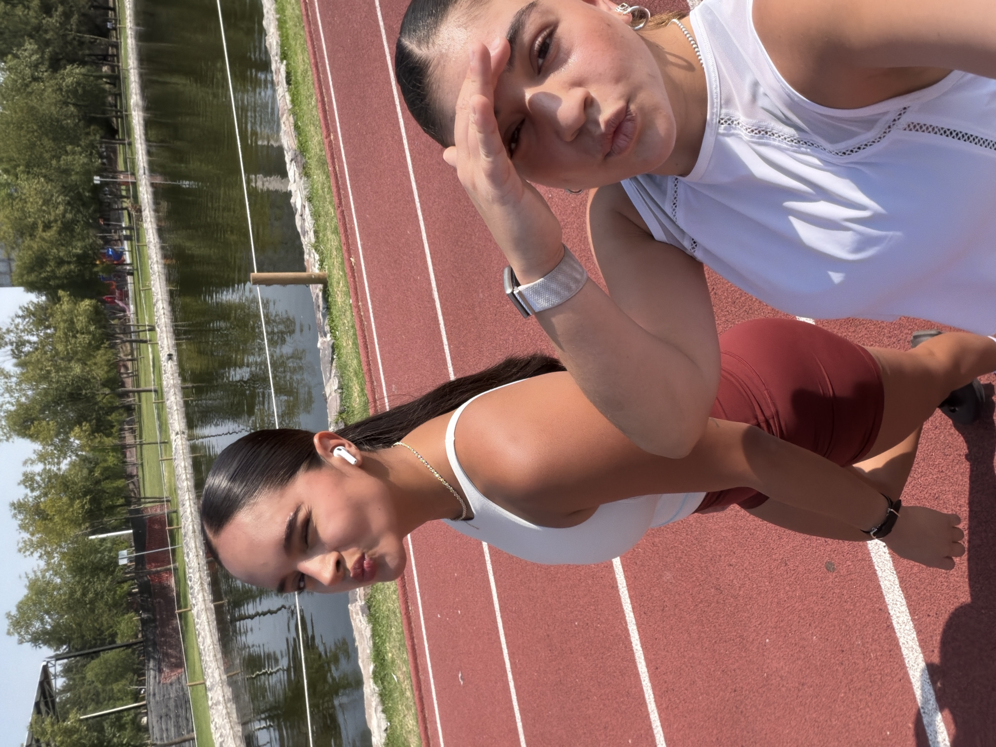 Two young women pose for a selfie on a red running track. Behind them is a pond with grassy banks and trees. The photo was taken in the Parque del Arte, in Puebla, Mexico. The woman closer to the camera is wearing a white tank top and maroon shorts. She has a silver chain necklace and a white earbud in her left ear. The other woman is wearing a white tank top and has a silver watch on her left wrist. Both women are puckering their lips towards the camera.