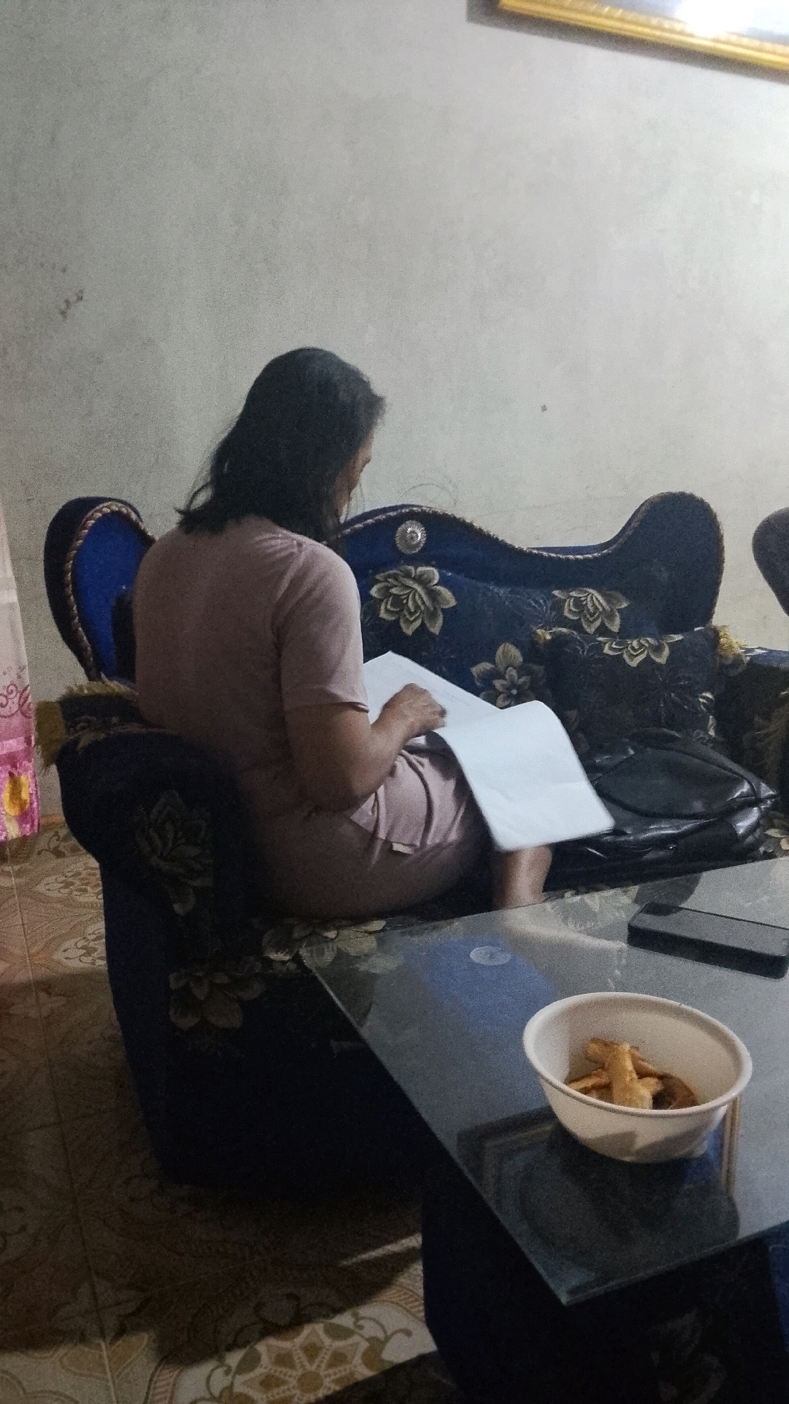 A woman is sitting on a patterned sofa, reading a document. She is wearing a light pink t-shirt and shorts. The sofa is dark blue with floral accents. In front of the sofa is a glass-top coffee table with a white bowl of snacks and a black mobile phone on it. The floor is tiled with a floral pattern. The walls are a neutral color. There is a framed picture on the wall in the upper right corner. The scene is likely indoors, in a residential setting.