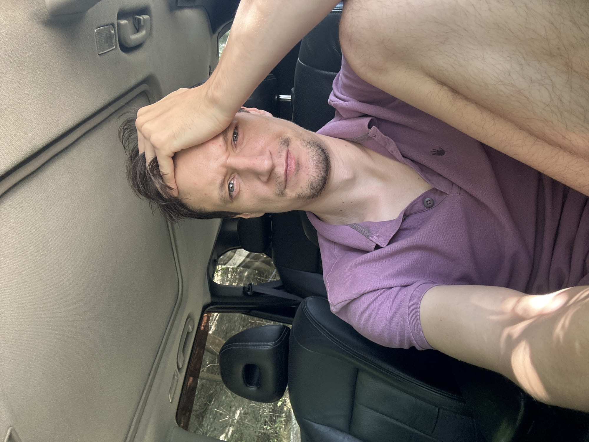 A man with short brown hair and a goatee is sitting in the driver's seat of a car, wearing a purple polo shirt. He is resting his right hand on his forehead and looking directly at the camera with a pensive expression. The interior of the car is visible, including black leather seats and a sunroof. The background outside the car windows shows trees and foliage, suggesting a rural or wooded area. The lighting indicates it is daytime, with sunlight casting shadows on the man's arm. There is no discernible indication of the city of Makiv, Ukraine, or any specific urban elements in the image.