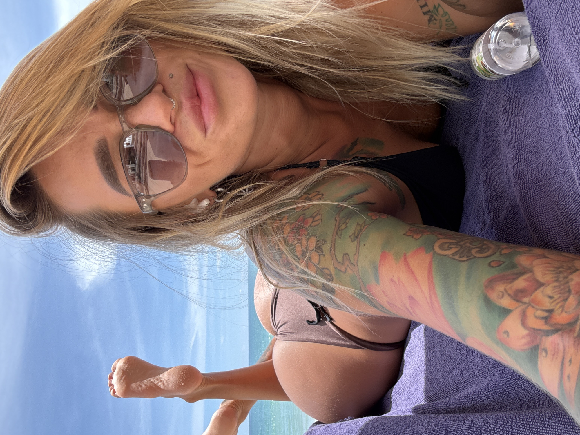 A fair-skinned woman with blonde hair is lying on her stomach on a purple towel on a beach in Choeng Thale, Thailand. She is wearing a dark bikini and sunglasses. Her left leg is extended upwards, with her foot visible. She has tattoos on her left arm and shoulder. The ocean and a blue sky with some clouds are visible in the background. A clear plastic bottle is on the towel next to her.