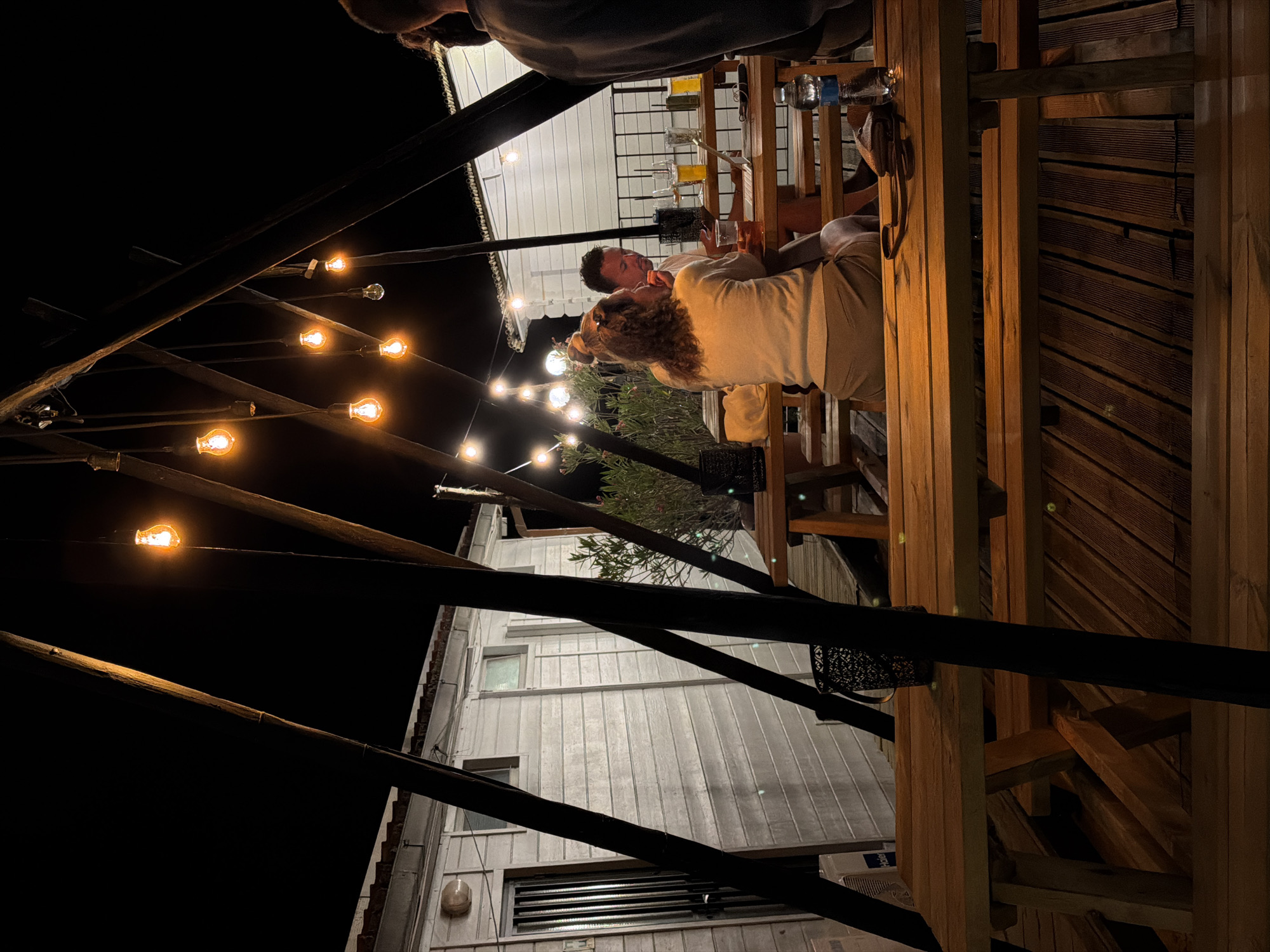 The image depicts an outdoor seating area at night, illuminated by string lights. In the foreground, a wooden picnic table and benches are visible. In the background, a white building with wooden siding and a door with a metal grate is present. Two individuals are seated at a table, looking at a laptop. Another person is partially visible on the right side of the image, with their back to the camera. There are several glasses of drinks on the table, as well as a bottle of water and a purse. The setting appears to be a casual outdoor dining or gathering space. The location is identified as Cavriglia, Italy.