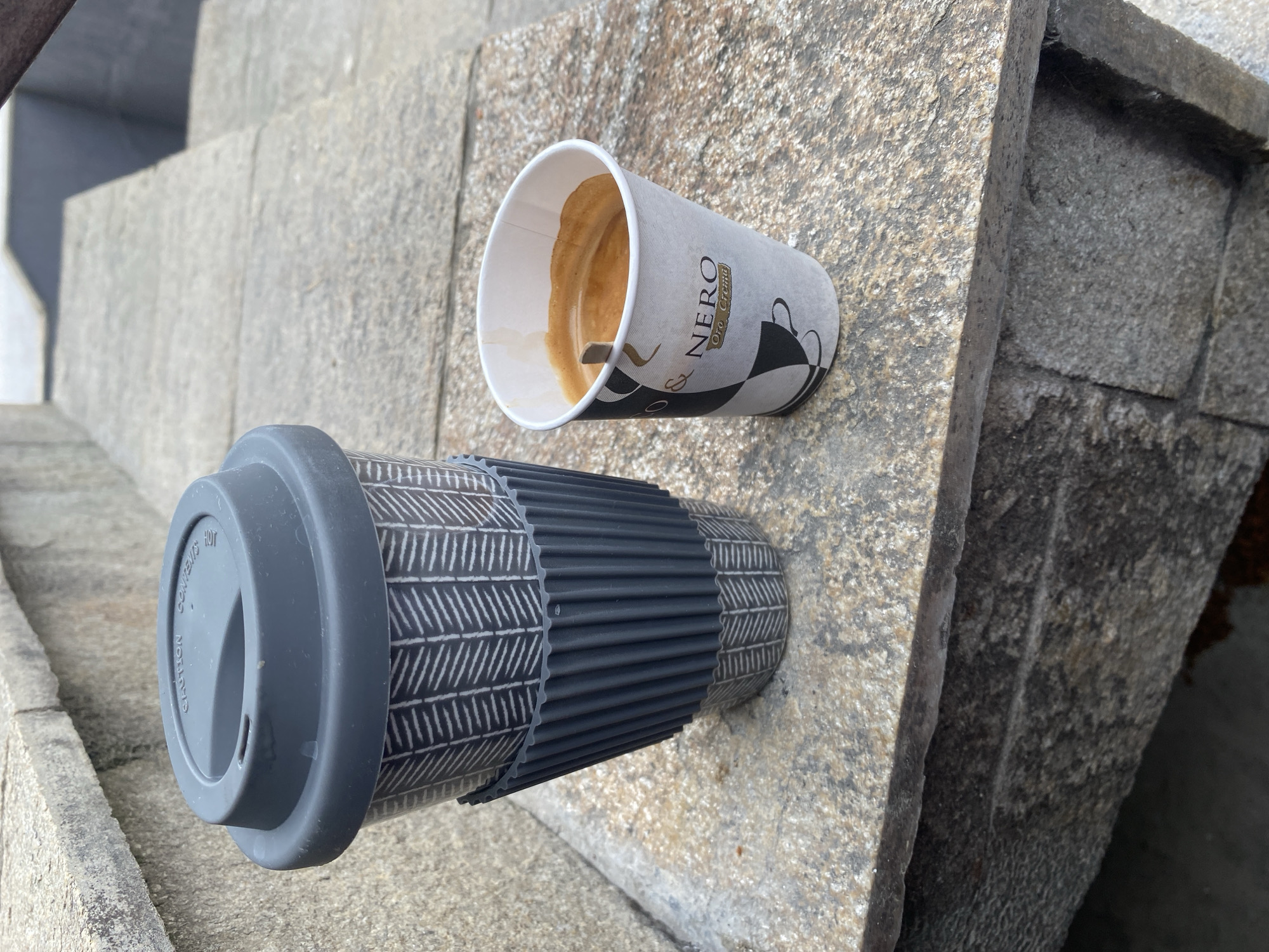 Two takeaway coffee cups are positioned on a stone surface, which appears to be part of an outdoor staircase or seating area. The cup on the left is a reusable travel mug with a grey lid and a dark grey ribbed sleeve over a patterned ceramic body. The cup on the right is a small paper cup with black and white geometric branding, containing a dark liquid, likely espresso, with a wooden stirrer inserted. The background shows further stone steps and the ground surface, which is also stone. The overall scene suggests a casual outdoor setting. The image is set in the city of Smolyan, Bulgaria.