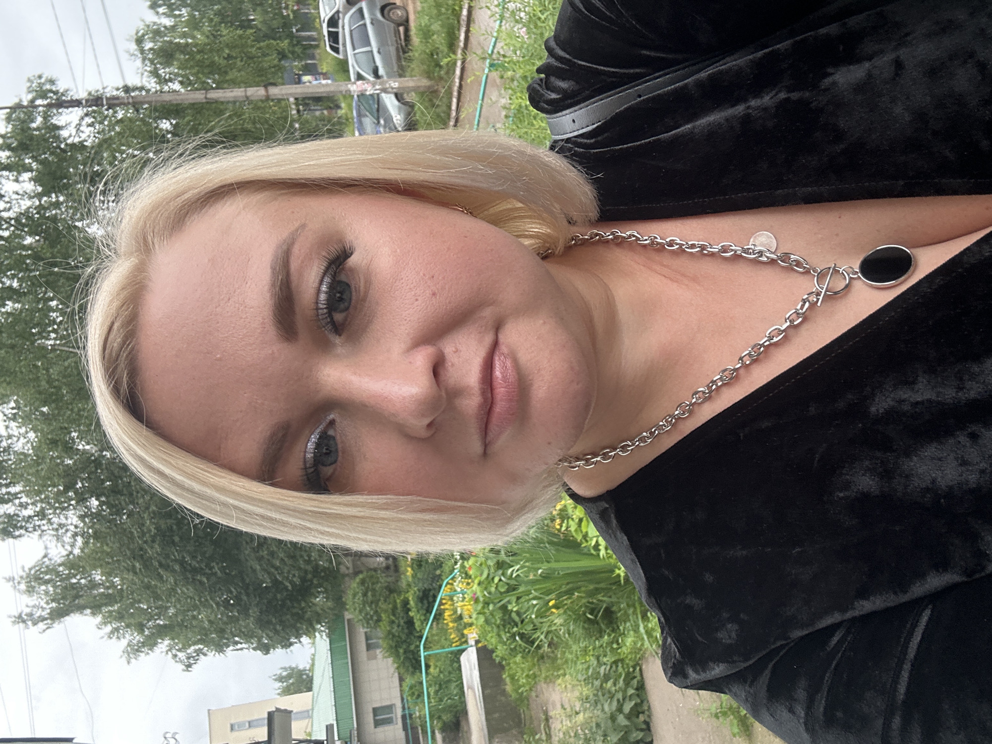 A woman with blonde hair and light skin is taking a selfie outdoors. She is wearing a black velvet top and a silver chain necklace with a black oval pendant. She has on silver eyeshadow and mascara. The background shows greenery, a building, and parked cars. The location is Chaikovsky, Russia, on a cloudy day.