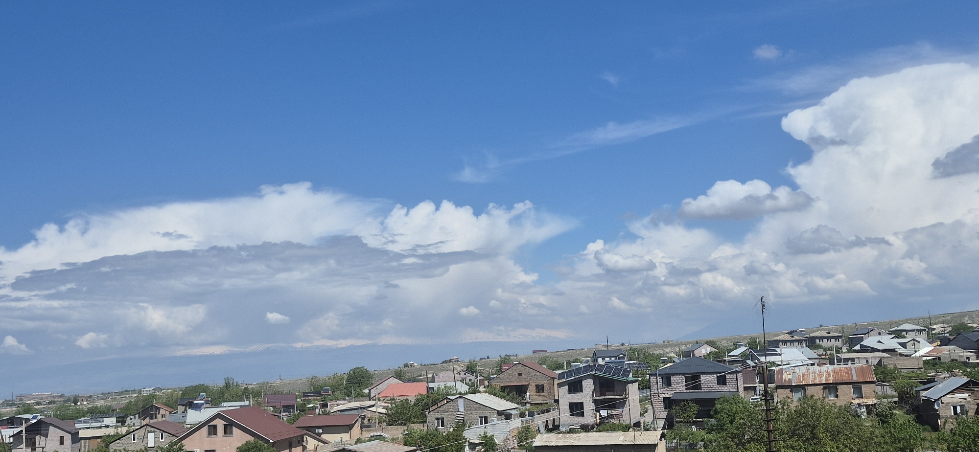 The image depicts a daytime view of a residential area, possibly a suburb, under a partly cloudy sky. The sky is predominantly blue with a scattering of white cumulus clouds. Below the sky, a cluster of houses with various architectural styles and roofing materials is visible. Some houses appear to have solar panels on their roofs. The houses are interspersed with trees and greenery. In the background, there is a faint outline of distant mountains, possibly snow-capped, suggesting a mountainous or high-altitude location. The scene is bathed in daylight, and the overall impression is that of a peaceful and settled residential environment.