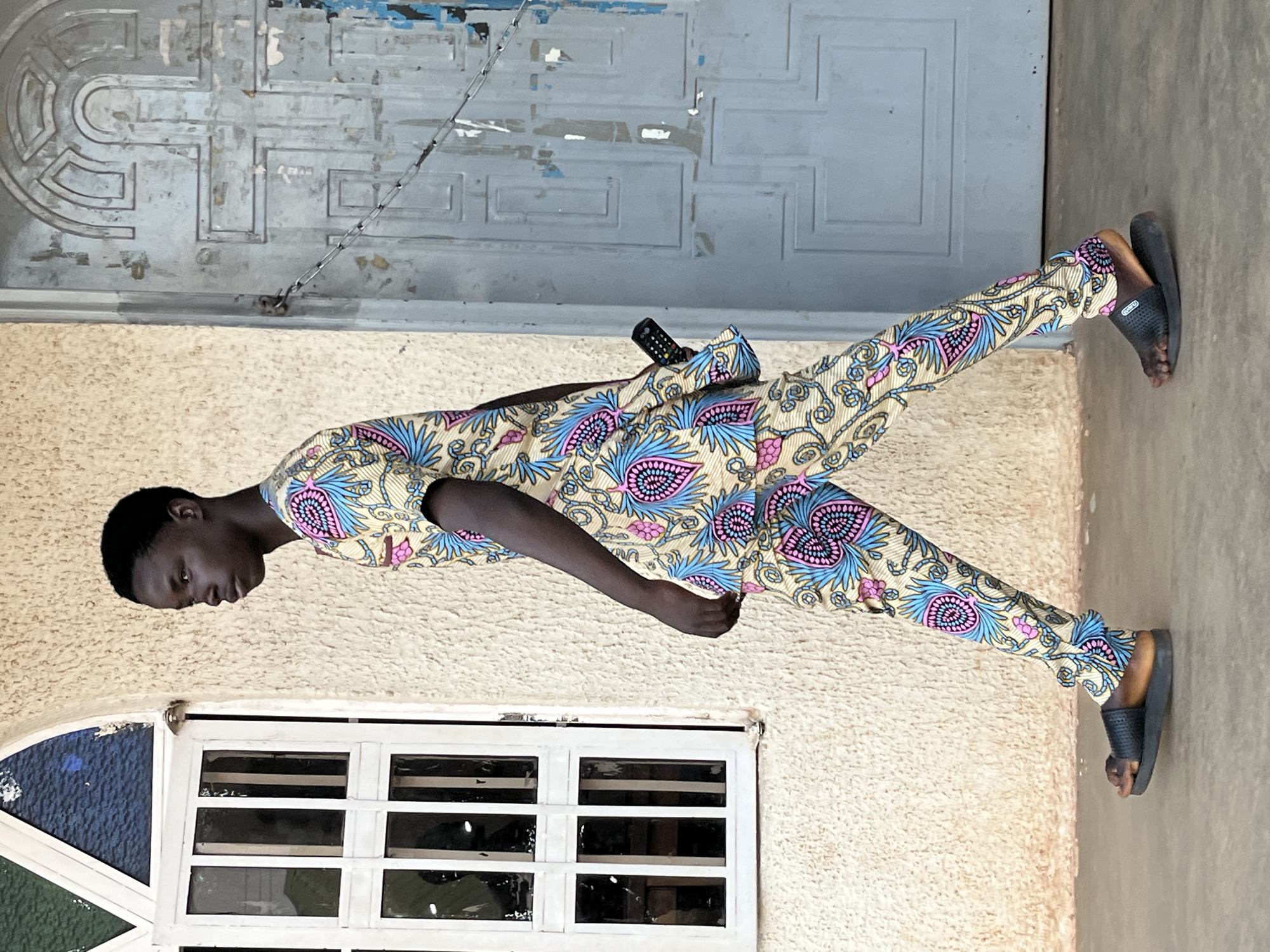 A person wearing a patterned beige suit with blue and pink floral designs stands against the exterior of a building. The background features a gray metal door above, with an off-white, textured wall below. A white-framed window with a blue accent is visible at the bottom. The person wears black sandals and has a small black remote control tucked into the suit. The location is plausibly within the city of Jos, Nigeria.