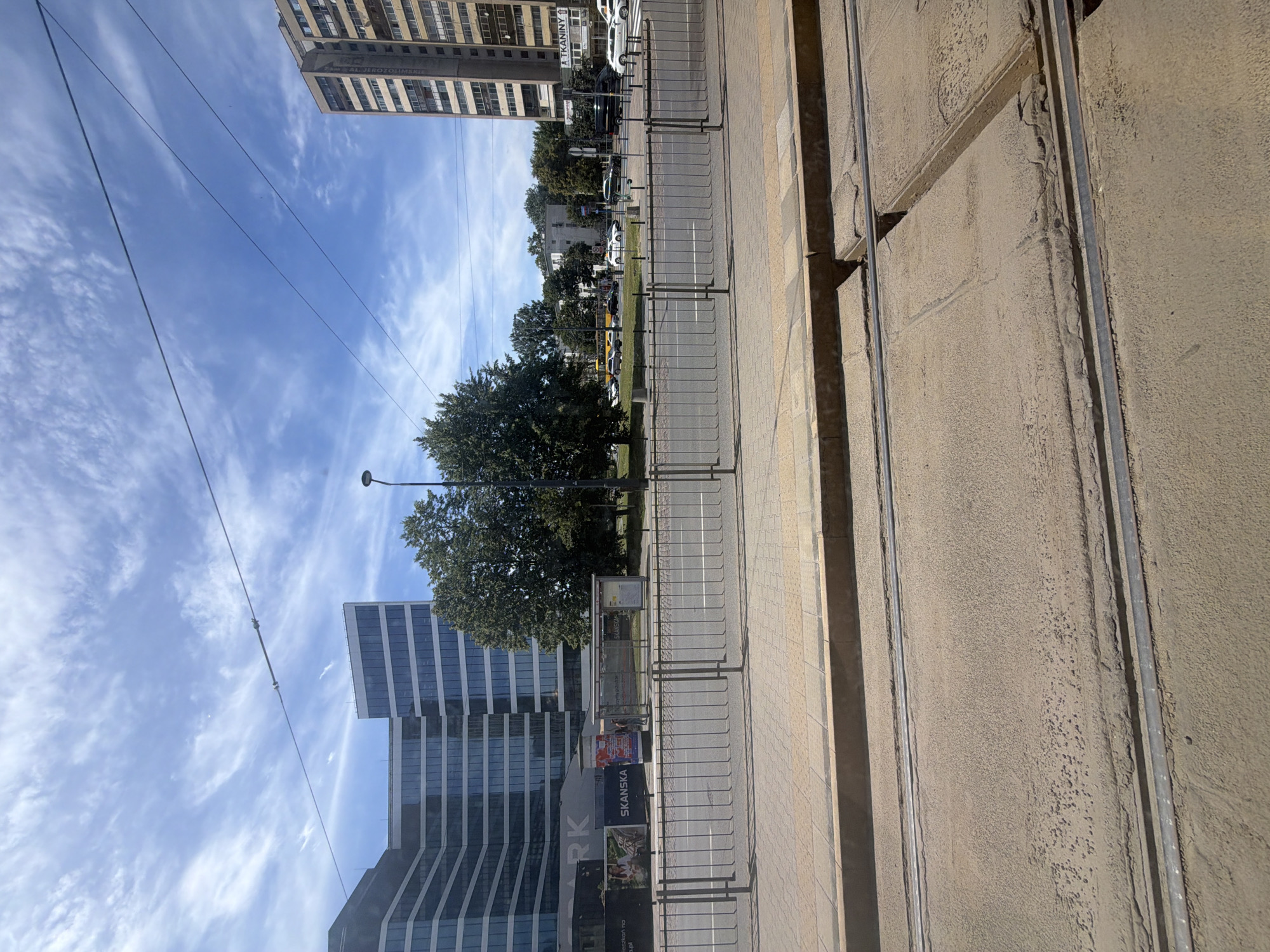 This is a daytime, eye-level, outdoor, wide-angle photograph taken from a ground-level perspective, likely from within a vehicle or near tram tracks. The foreground features tram tracks set into a dusty concrete surface, with the edges of the tracks and surrounding pavement visible. A metal railing separates this area from a sidewalk and a street.
In the mid-ground, across the street, are several elements. A bus stop shelter is present, with a single person standing inside. Behind the bus stop, a modern, multi-story office building with a glass facade dominates the left side of the frame. To the right of the office building, a cluster of mature trees stands, followed by a tall, older, tan-colored apartment building on the far right, adorned with visible windows and some signage. The street itself is visible with several cars and a yellow bus on it, suggesting moderate traffic. The sky above is a bright blue with scattered white clouds, and overhead power lines for the tram are visible. The overall impression is of an urban environment with a mix of modern and older architecture, likely during fair weather.
