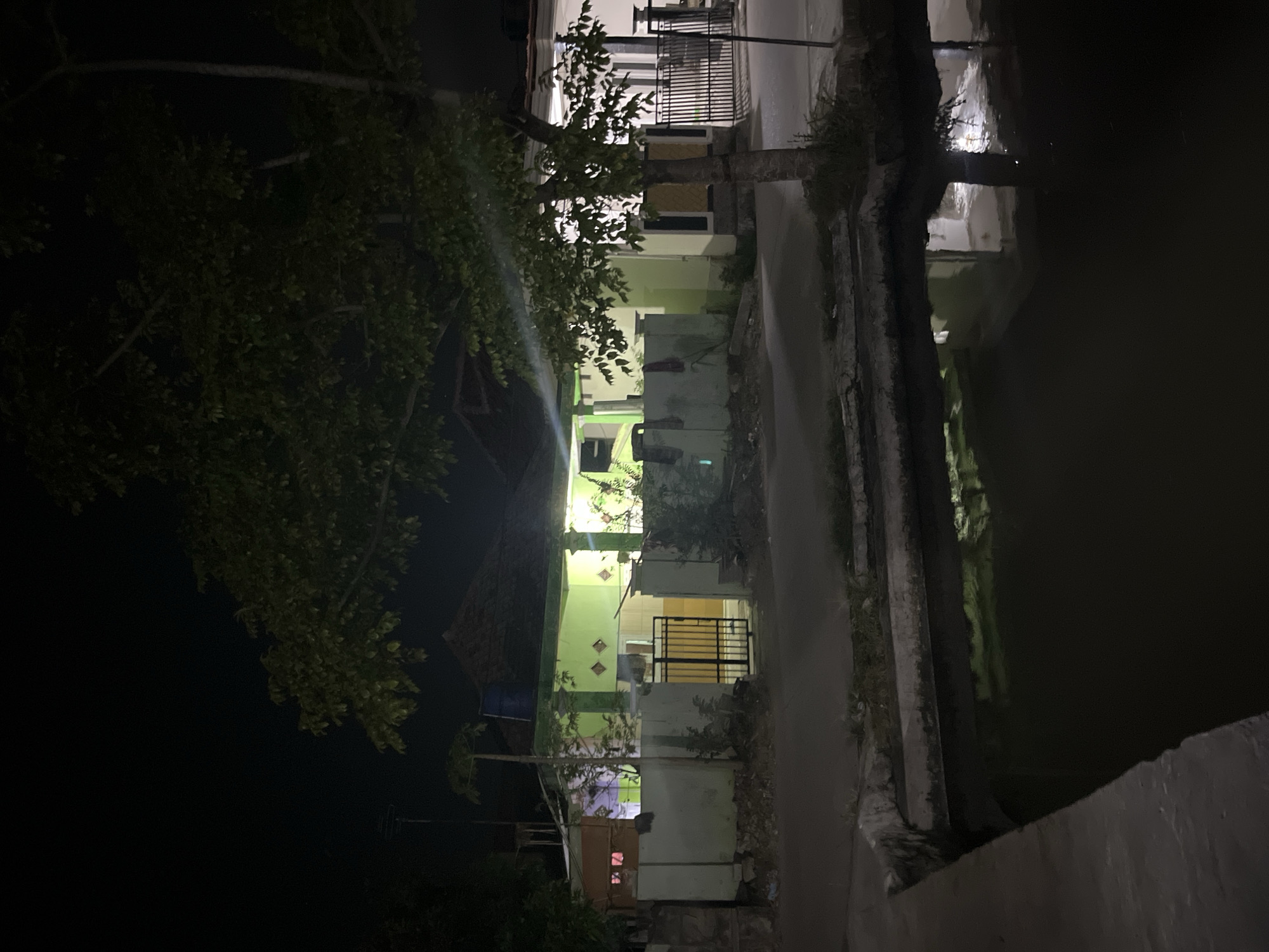 The image depicts a residential area at night in Laban, Indonesia. In the foreground, a dark body of water with a concrete edge reflects the lights from the houses. Behind the water, a paved road separates it from a row of houses and fences. The houses are illuminated from within, casting a green glow on some of their facades. A metal gate is visible in front of one of the houses. Trees with dark green foliage are scattered throughout the scene, with a prominent tree in the mid-ground. The overall impression is of a quiet, residential street at night.