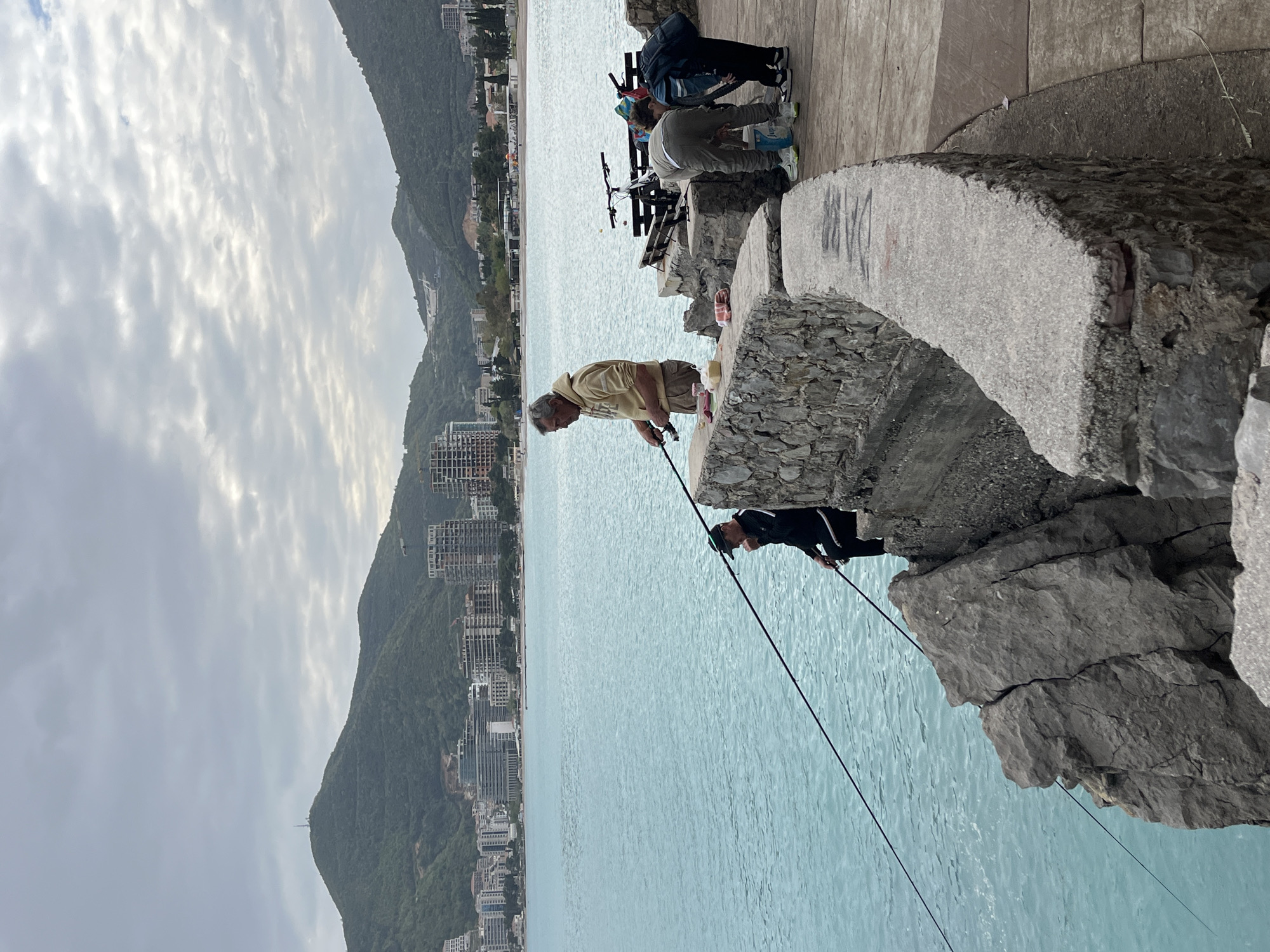 A horizontal, eye-level shot depicts a group of individuals fishing along the Budva, Montenegro coastline. Four figures are observable, two of whom are actively fishing, one in a tan jacket and the other in a black jacket, with fishing poles in hand. Buildings in the background on the right, a mountain on the left, and a stone wall create a pier. The sky is heavily overcast.