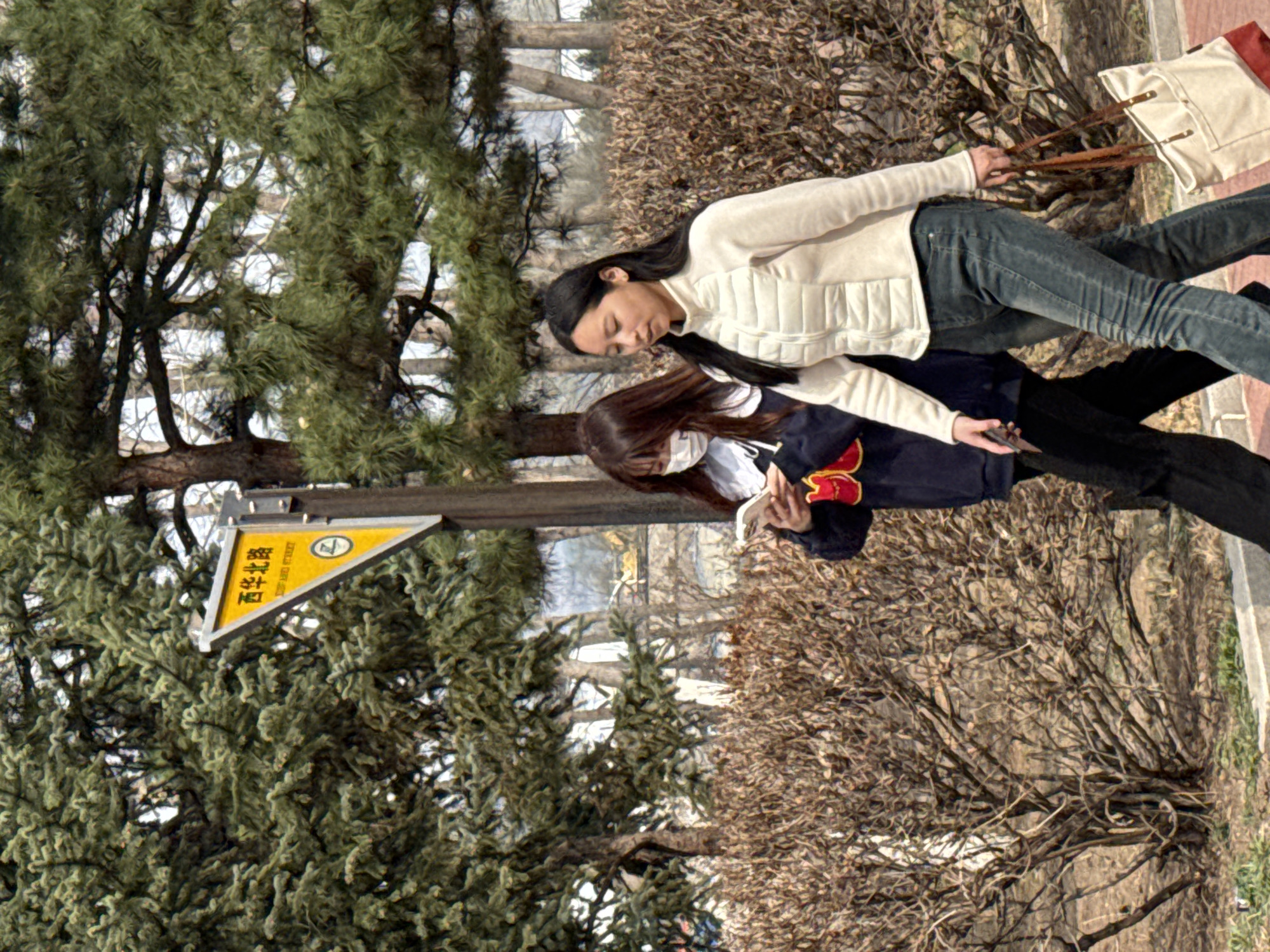 Two women are walking on a sidewalk next to trees and bushes. The woman closest to the viewer is wearing a white vest, jeans, and holding a bag with sticks inside. The other woman is wearing a dark blue jacket with a red design, black pants, and is looking at a phone. A street sign is visible on the left side of the image.