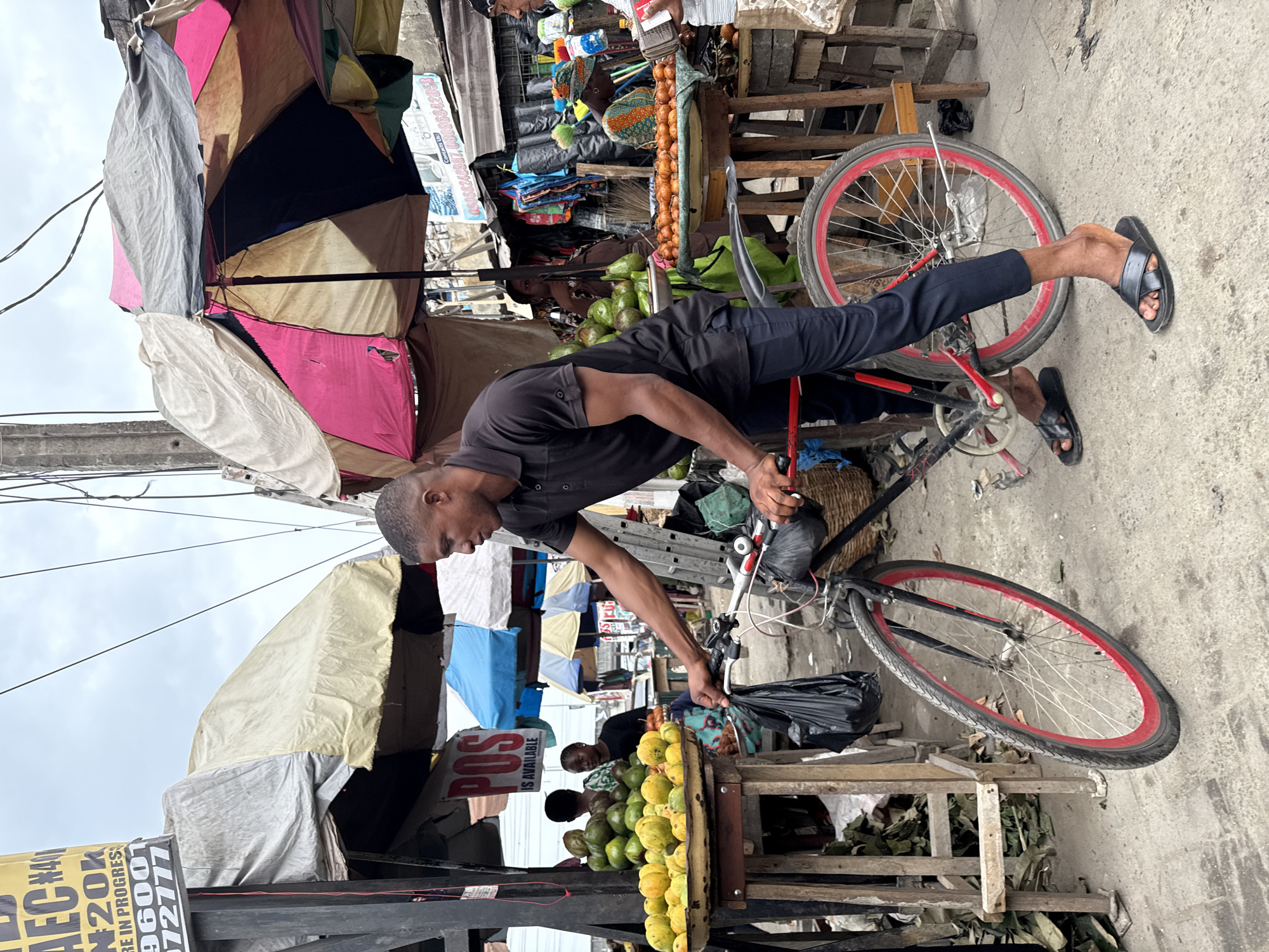 The image depicts a street scene in Lagos, Nigeria, likely a market area. A man, dressed in black, is precariously balanced on a red and black bicycle. He is standing on the seat with one foot on the back wheel and appears to be loading a black plastic bag onto the handlebars. Around him are stalls with fruits, possibly avocados and mangoes, displayed for sale. Other people are visible in the background, suggesting a busy environment. Overhead, a collection of umbrellas provides shade, adding to the cluttered and vibrant atmosphere of the scene. Signage is visible with some text, including "POS is AVAILABLE".