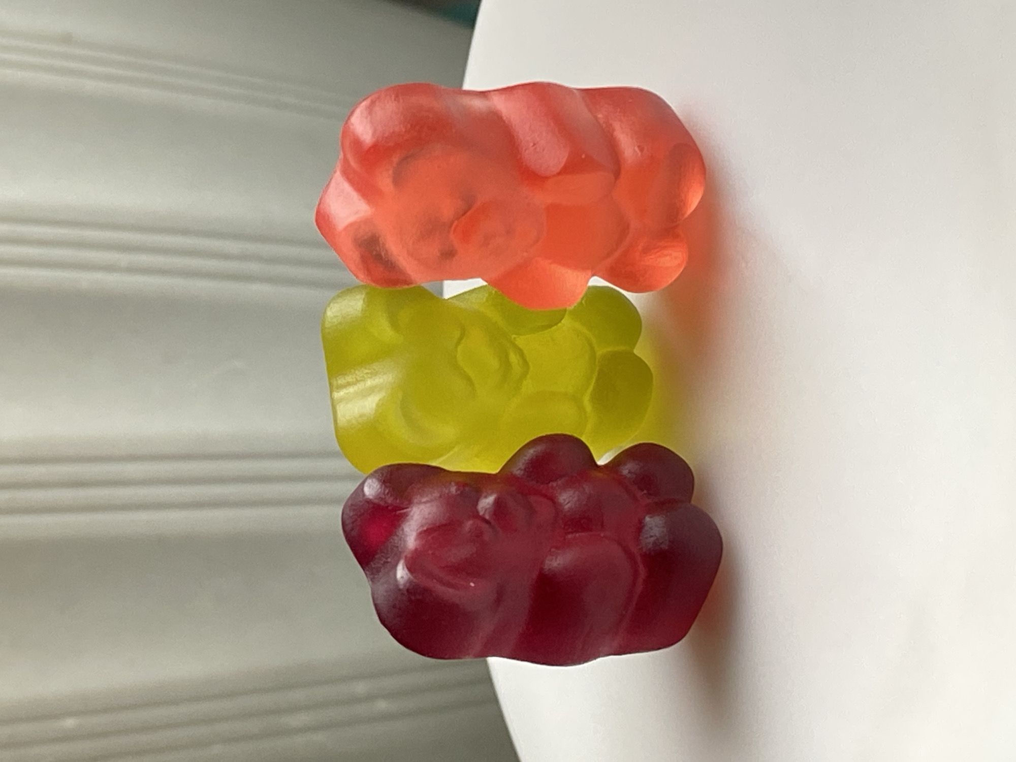 The image features three large, translucent gummy candies arranged in a vertical stack or line. From top to bottom, the gummies are red-orange, yellow-green, and deep red-purple. Each gummy has a distinct, somewhat irregular and textured shape, giving them a sculpted appearance rather than a smooth finish.
The background on the left side of the image is a light grey or off-white surface with faint, horizontal lines or grooves, possibly a wall panel or a textured board. The right side of the background is a plain, solid white surface, creating a clean, contrasting backdrop. The lighting is bright and even, emphasizing the translucent quality and vibrant colors of the gummies. No specific activity is taking place beyond the static arrangement of the candies. There are no people, animals, or visible text in the image. The scene appears to be an indoor, close-up still life.