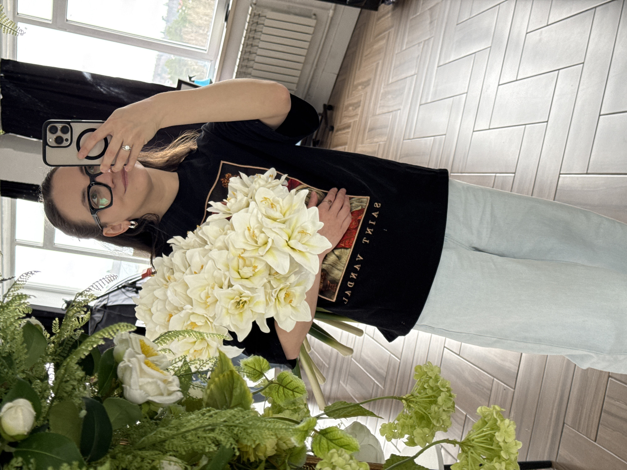 This mirror selfie, taken in Chaikovsky, Russia, captures a woman holding a large bouquet of white flowers with yellow centers. The woman, with dark brown hair pulled back, wears black-rimmed glasses and has a neutral to slightly pleasant expression. She is dressed in a black short-sleeved t-shirt featuring a mirrored graphic design resembling classical art and part of the text "ГАЯТ ТИАS" (likely "SAINT YAGAN" when un-mirrored), paired with light-colored jeans. A silver ring is visible on her left ring finger. She holds an iPhone, equipped with a light-colored case and a black MagSafe ring, to capture her reflection.
The indoor setting is brightly lit by natural light streaming through a large window, behind which a blurred outdoor environment is visible. A white radiator is present near the window. The floor exhibits a light-colored, herringbone pattern, possibly of wood or tile. In the foreground, below the main bouquet, a decorative arrangement of artificial greenery, including ferns, light green hydrangea-like flowers, and additional white flowers, adds to the scene. The image depicts a casual, daytime moment, possibly showcasing a new floral arrangement.