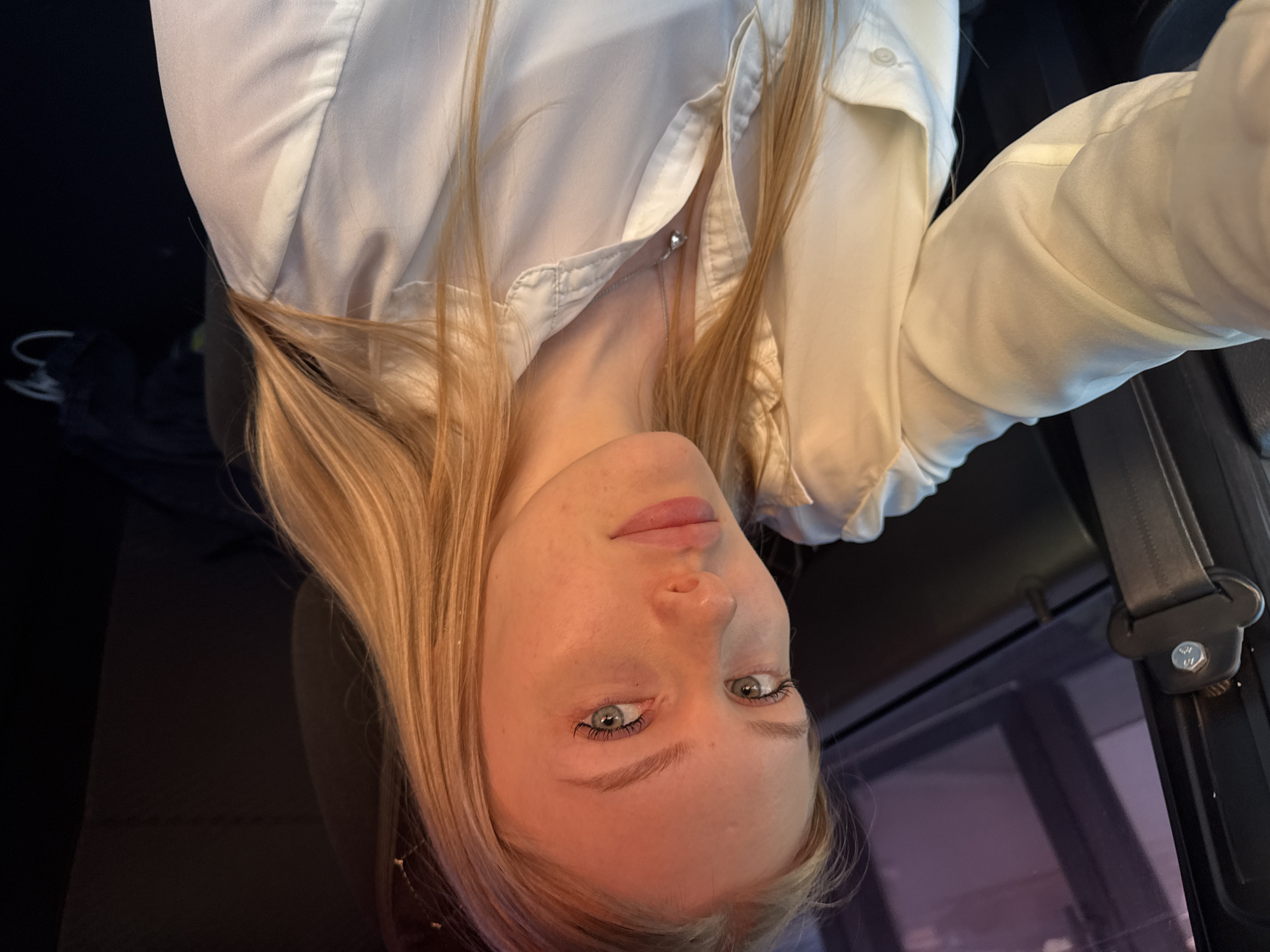 A fair-skinned woman with blonde hair is pictured from a low, tilted angle. She is wearing a white, button-down shirt and a silver necklace. She has green eyes and light eyebrows. The background shows a dark interior, possibly of a vehicle, with a seatbelt visible on the right side of the frame. The interior reflects a city scene at night with the purple-ish reflection of street lights. The location is potentially in a vehicle traveling at night. It is not possible to verify that it is the city of Perm, Russia.
