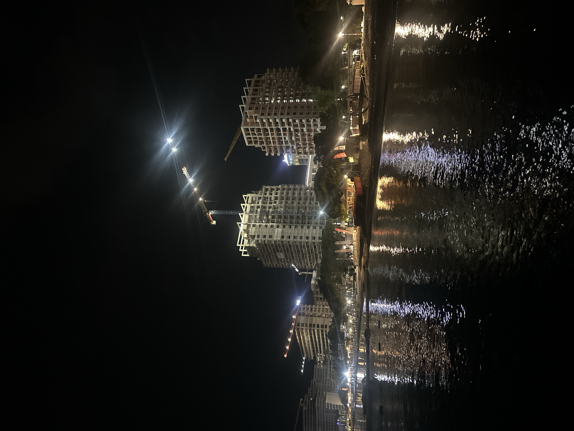 The image depicts a nighttime scene along the coast of Budva, Montenegro, looking from a body of water towards a developed shoreline. Multiple high-rise buildings, predominantly under construction, dominate the mid-ground. Several large tower cranes, some illuminated with red lights at their highest points, are positioned around or atop these structures. The two most prominent buildings in the center appear as concrete skeletons, revealing their internal floor and column frameworks, with some scaffolding visible on the taller one to the right.
The shoreline between the buildings and the water features a strip of land, likely a beach or promenade, extensively illuminated by numerous streetlights and light sources from smaller structures, possibly kiosks or temporary businesses. Red objects, potentially boats or beach equipment, are visible on the sand to the left. A wooden walkway or pier-like structure extends into the water on the right.
The foreground is a dark body of water, likely the Adriatic Sea, which vividly reflects the intense artificial lights from the shore and buildings. These reflections appear as elongated, shimmering streaks of white, yellow, and orange across the subtly rippled water surface. The sky above is deep black, with several bright, star-like light sources exhibiting distinct lens flares in the upper right quadrant, likely originating from distant cranes or powerful spotlights. No individual people are distinctly discernible within the frame, but the extensive illumination and infrastructure indicate a lively urban coastal environment.