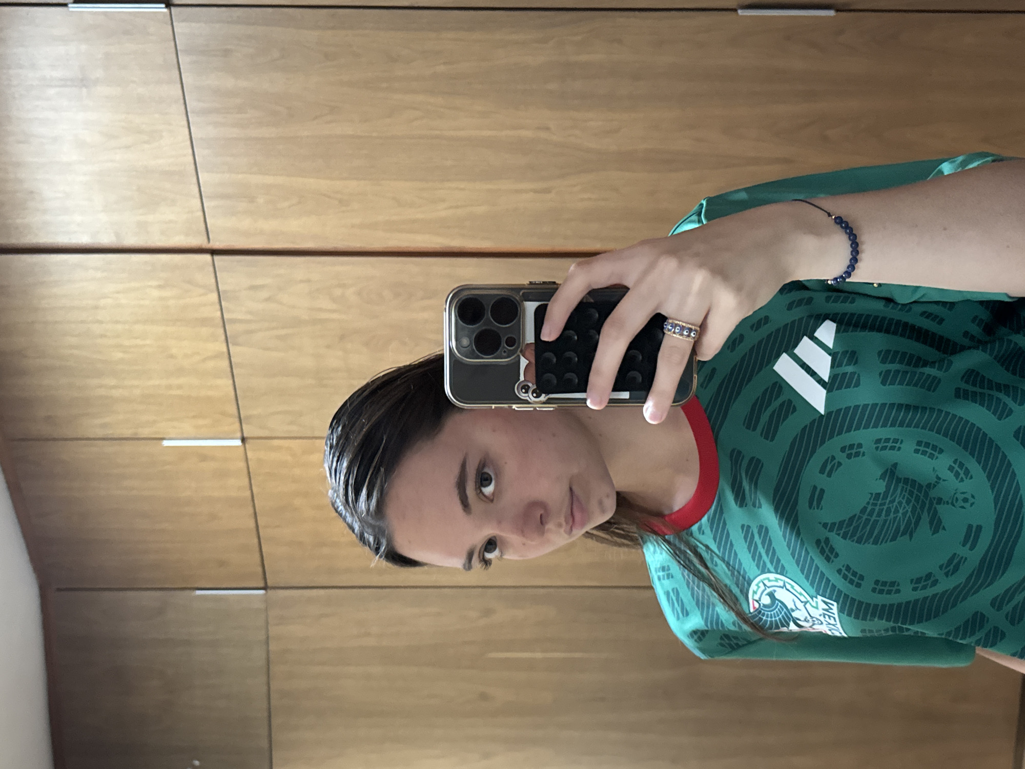 A person is taking a selfie in front of a wooden closet. The person is wearing a green Mexico soccer jersey. The phone is a dark color, and the person is wearing a ring on one finger and a bracelet on their wrist. The location is not clear from this image and it is not possible to verify if this image was taken in San Bernardino Tlaxcalancingo, Mexico.