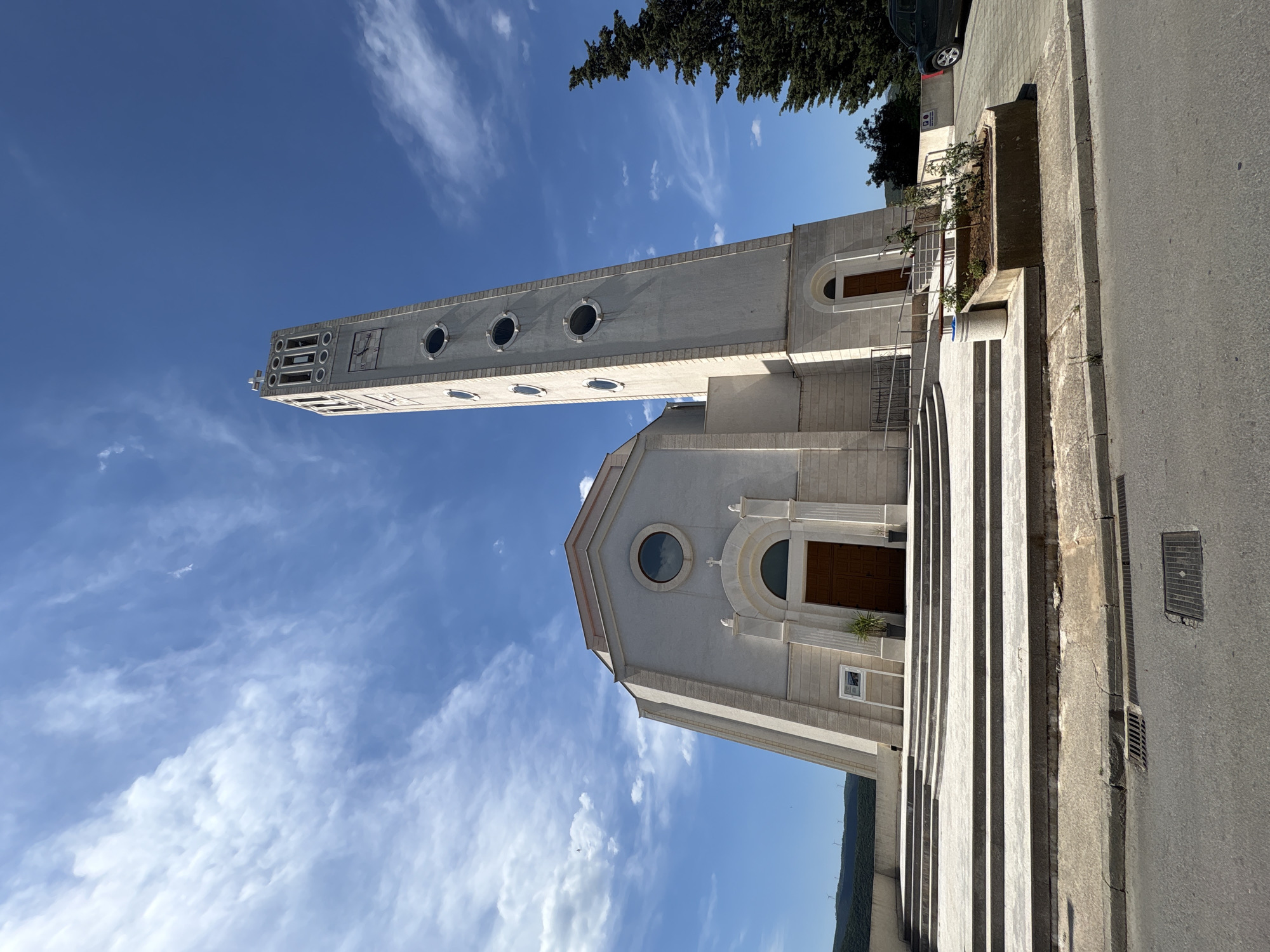 The image shows the Church of Christ the King in Neum, Bosnia and Herzegovina, viewed from a low angle. The church is a light-colored building with a tall, slender bell tower featuring circular windows. The facade has a large arched doorway and a circular window above. A wide set of steps leads up to the entrance. The sky is blue with scattered white clouds. A road and some greenery are visible on the right side of the frame. A tree is visible partially obscuring the church from the right.