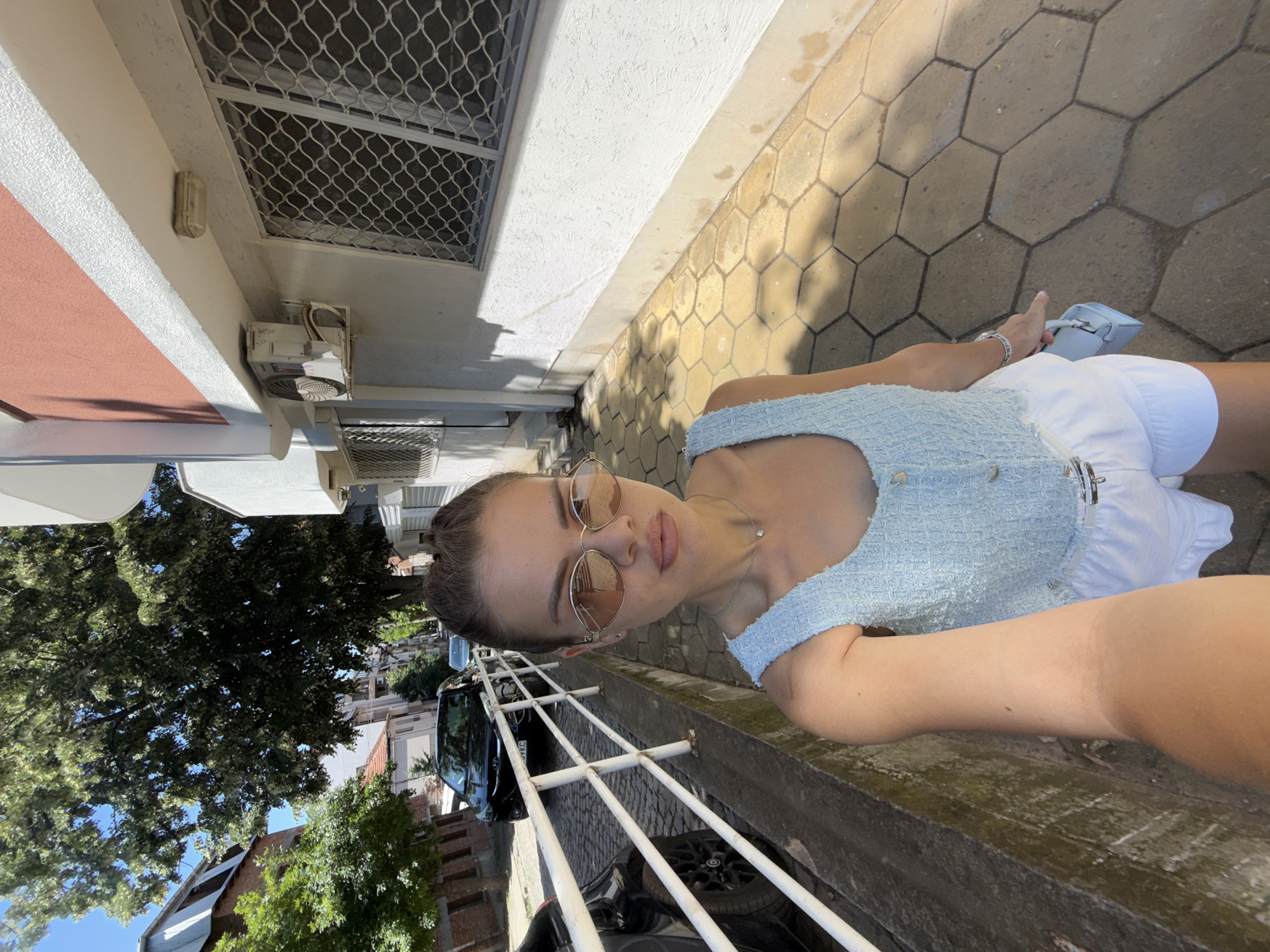 A woman with brown hair pulled back stands on a hexagonal-tiled sidewalk next to a white metal railing. She is wearing a light blue textured sleeveless top, white ruffled shorts, and oval-shaped sunglasses. She is holding a small light blue handbag in her right hand and her left arm is extended towards the camera, suggesting she is taking a selfie. Behind her, a street is visible with parked cars on a cobblestone surface. Buildings line the street, one of which is a light-colored structure with a pink upper section and visible air conditioning units and barred windows. Trees with green foliage are also present. The scene appears to be a street in Plovdiv, Bulgaria, under a clear blue sky.