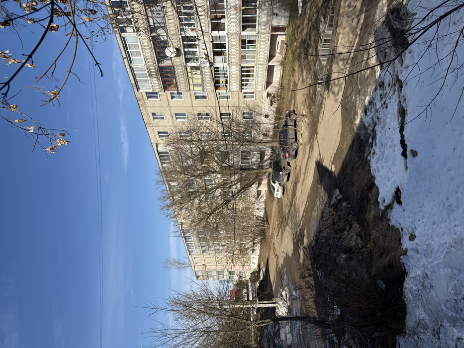 This image depicts an urban residential scene in Kurganovo, Russia, under a clear blue sky. A large, multi-story apartment building with a light-colored facade, featuring numerous windows and enclosed balconies, dominates the middle and right sections of the frame. One balcony on an upper floor displays a satellite dish. The ground level comprises an unpaved dirt and gravel area, serving as a parking lot, with visible patches of melting snow, mud, and water puddles. In the immediate foreground, a substantial mound of dirty, melting snow and earth is present. Three soiled passenger vehicles are parked along the apartment building: a white sedan, a dark-colored sedan, and another dark-colored sedan. Numerous deciduous trees, all bare of leaves, are scattered throughout the scene, with their branches partially obscuring the building and sky. Bare bushes are visible near the building's base. Overhead power lines traverse the upper portion of the image. The scene is illuminated by direct sunlight, casting distinct shadows. No individuals or discernible activities are visible.