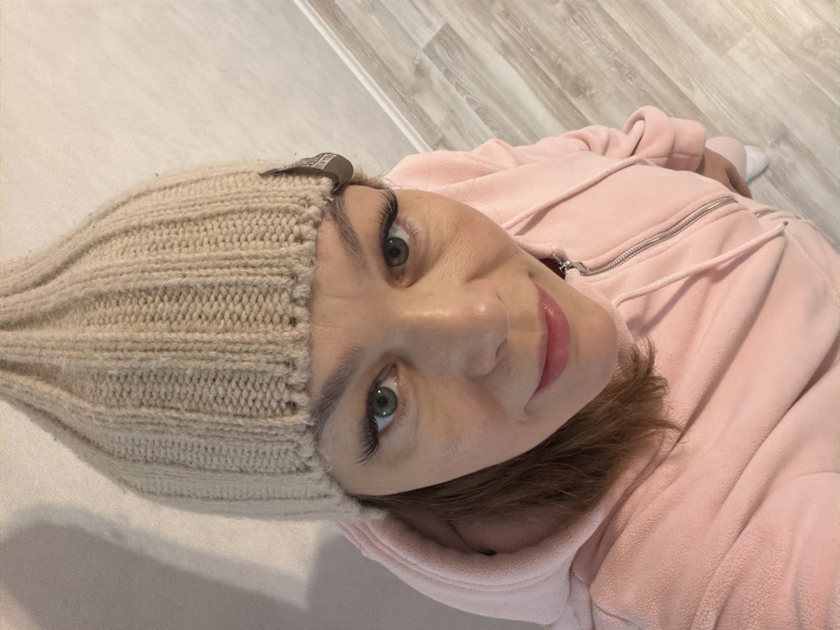 The image captures a close-up, high-angle view of a person, likely female, lying down, looking towards the camera. The image is rotated 90 degrees counter-clockwise, showing the person's head on the left and body extending to the right.
The person is wearing a light beige or cream-colored knitted beanie with a prominent ribbed texture. A small, brown rectangular tag with faint, unreadable text or a logo is attached to the beanie above the left eyebrow. The person's face is well-lit, revealing clear skin and a slight, gentle smile with pink, glossy lips. Her eyes are a striking green color, framed by long, dark eyelashes (possibly extensions) and neatly shaped eyebrows. Brown hair is visible peeking out from under the beanie and around the neck area, contrasting with the light-colored headwear.
She is dressed in a light pink, possibly fleece or velour, zippered hoodie or jacket. The silver zipper is visible down the center of the garment, and light pink drawstrings are also present around the hood opening. The hood appears to be up or at least surrounding her head.
The background consists of a light-colored surface. To the left, there is a solid, lighter beige or off-white surface, possibly a wall or a different floor section. To the right and behind the person, a light wood or laminate floor with visible plank lines and a subtle grain pattern is present. In the upper right corner of the image, a small portion of what appears to be a white sock or footwear is visible, suggesting her legs extend further into the background.
The overall impression is one of relaxation, possibly a casual selfie taken indoors. The lighting is bright and even, without harsh shadows, suggesting an indoor setting with good artificial or natural light.