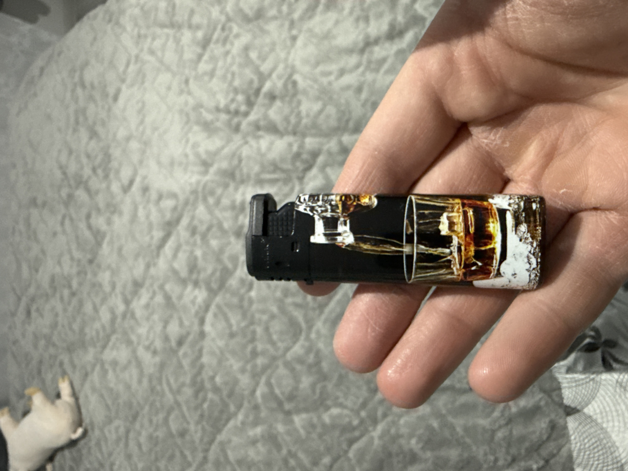 An indoor close-up shot captured in Stara Zagora, Bulgaria, shows an adult's hand holding a black cigarette lighter. The lighter features a distinctive design that mimics a glass filled with an amber liquid, possibly whiskey or a similar spirit, complete with ice cubes. The design wraps around the body of the lighter, creating an optical illusion that makes it appear like a miniature drink. The hand is positioned horizontally, with fingers gently wrapped around the lighter, and shows visible skin texture. The background consists of a light-colored, possibly off-white or light gray, quilted or textured fabric, likely a bedspread or blanket, which appears slightly out of focus. In the bottom-left corner of the frame, a blurry, small, light-colored object, possibly a toy pig, is partially visible lying on the textured surface. The lighting appears to be artificial, casting soft shadows. No discernible text is visible on the lighter or in the surroundings.