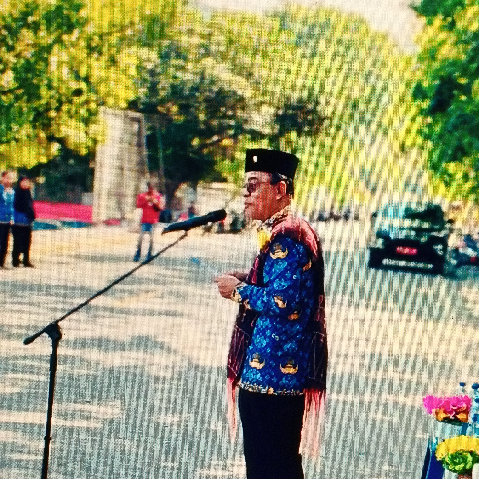 The image shows a man in traditional Indonesian attire, including a black cap and a blue patterned jacket with gold emblems, speaking at a microphone. He is wearing sunglasses and pointing with his right hand. Behind him, the background is blurred, but it appears to be an outdoor setting with trees and a road. Several other people are visible in the background, some wearing casual clothing and others in what appears to be formal wear. A black car is parked on the road in the mid-ground. The scene suggests a public event or speech taking place in the city of Ende, Indonesia.