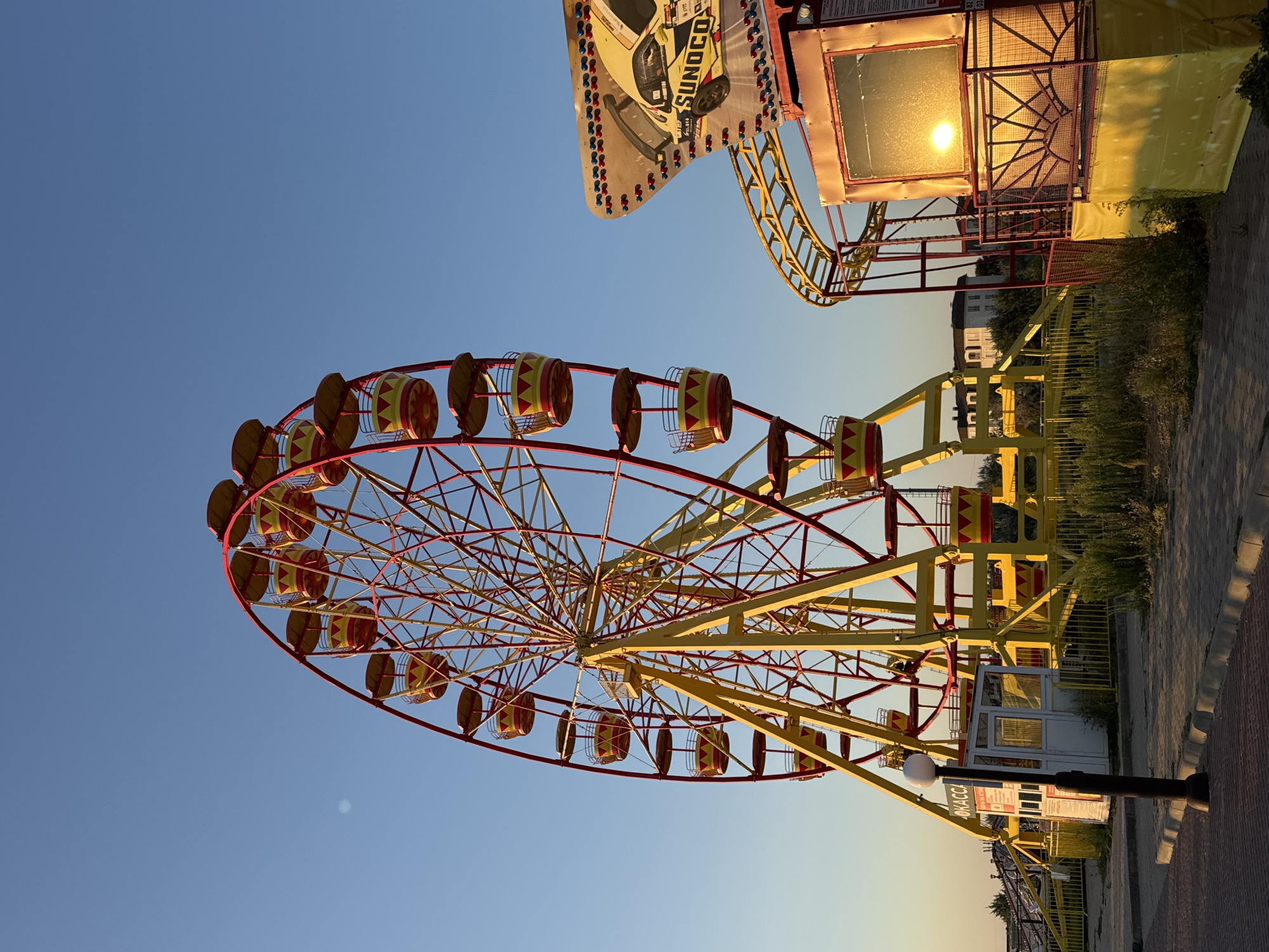 An amusement park is shown on a sunny day in Novofedorovka, Ukraine. In the foreground, a Ferris wheel with yellow and red gondolas is on the left, and a roller coaster with yellow tracks and red supports is on the right. The Ferris wheel is tall and wide, with many gondolas. The roller coaster has a loop and a sharp turn. In the background, there are some buildings and trees. The sky is clear blue, and the sun is shining brightly. There are no people visible in the image.