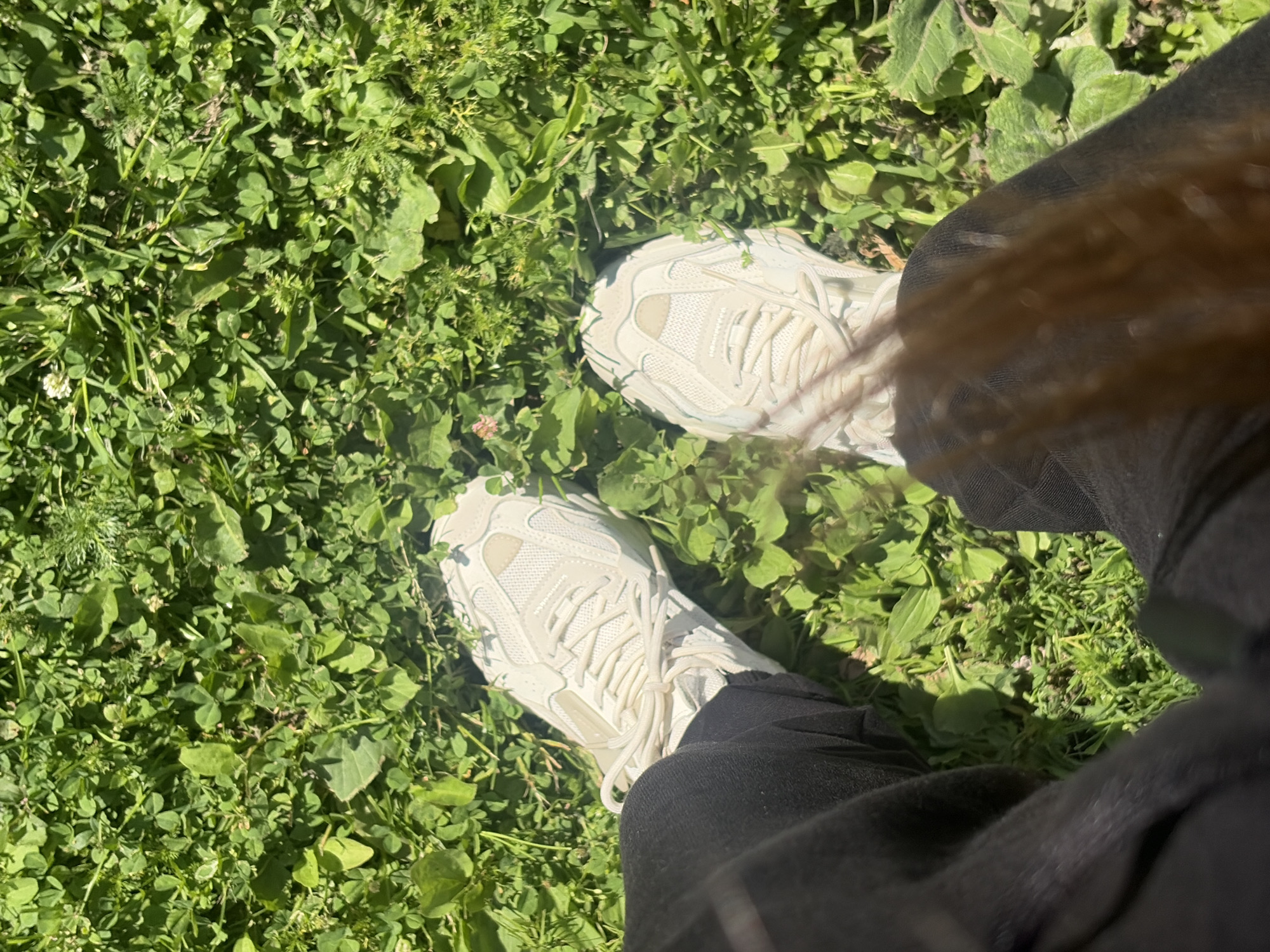 The image shows a person's legs and feet, wearing white athletic shoes with thick soles and dark pants. The person is standing in a patch of lush green grass, with some clover and other small plants visible. The sun is shining, casting dappled shadows on the ground, suggesting it is daytime, likely morning or afternoon given the bright lighting. The setting appears to be an outdoor green space, possibly a park or garden. A strand of blonde hair is also visible in the foreground, obscuring part of the shoes and pants. No text is visible in the image.