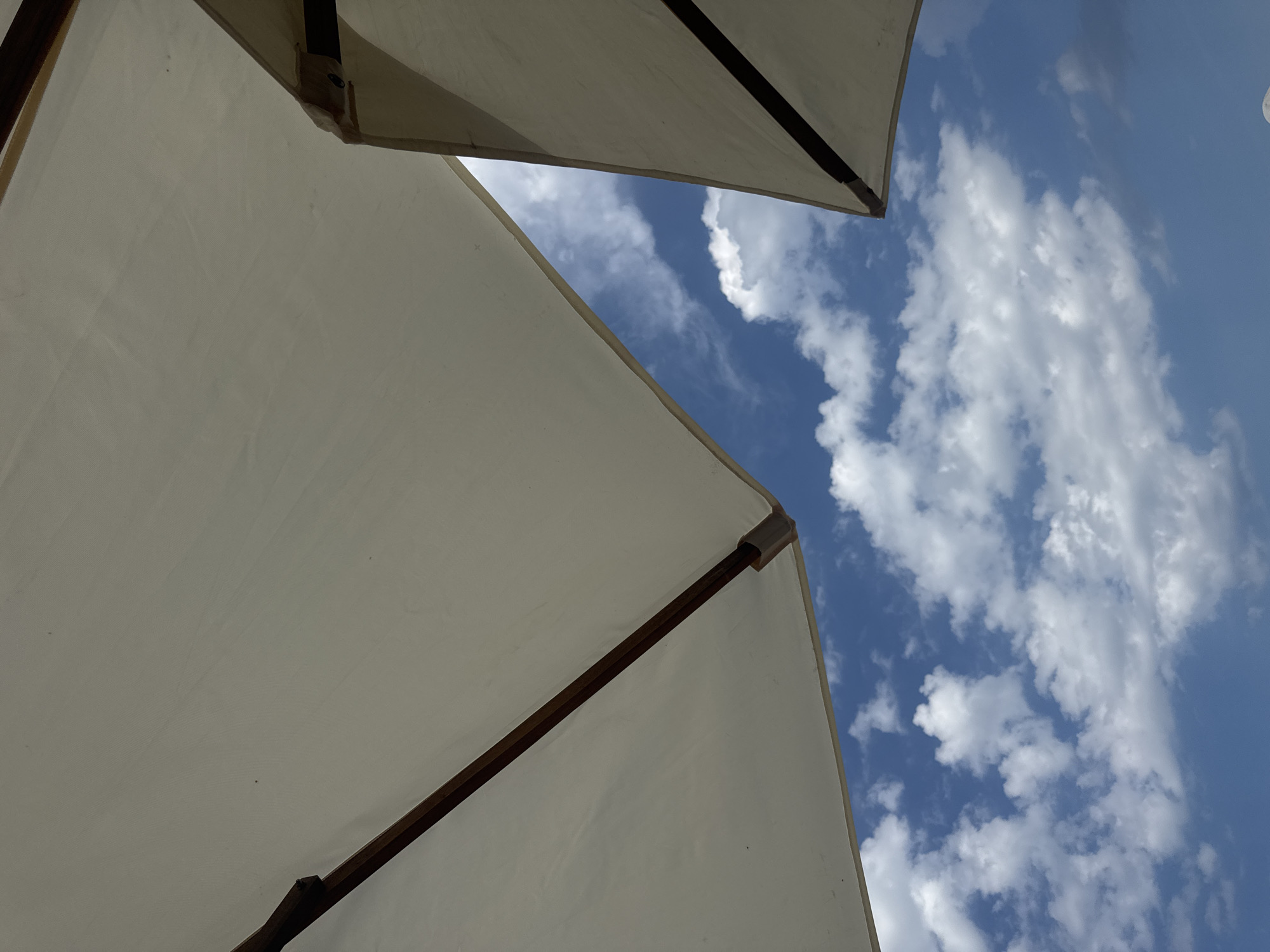 The image shows an upward view of two cream-colored umbrellas against a bright blue sky with scattered white clouds. The umbrellas are positioned in the foreground, with their fabric taut and supported by wooden frames. One umbrella is larger and takes up the majority of the left side of the frame, while a portion of a second, smaller umbrella is visible in the upper right. The sun is not directly visible, but the bright sky and clouds suggest it is daytime, likely late morning or afternoon, given the clear visibility of the clouds. There are no people or specific landmarks visible. The scene appears to be an outdoor setting, possibly a beach, resort, or cafe, where umbrellas are used for shade. The overall impression is one of a pleasant, sunny day. No text is visible in the image. The location context of Nha Trang, Vietnam, implies a warm, tropical climate.
