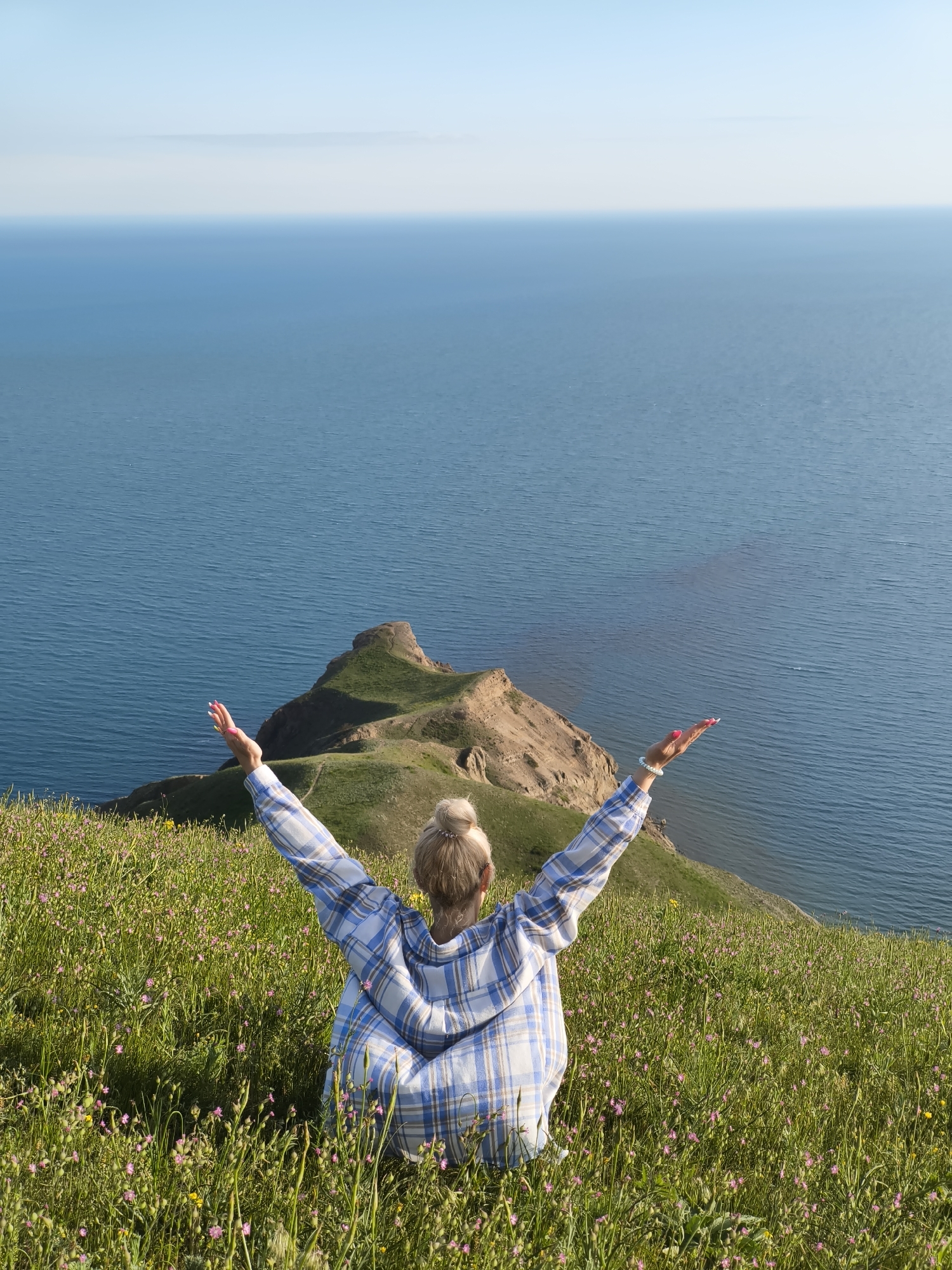 A full, high-angle shot depicts a woman seated on a grassy hillside, facing away from the viewer, with her arms raised in a posture of apparent joy or exhilaration. The scene is set in Коктебельский поселковый совет, Ukraine.
The woman, who appears to be Caucasian, has blonde hair pulled back in a bun. She is wearing a light blue and white plaid shirt or jacket. The hillside is covered in green grass and dotted with small wildflowers, primarily pink and yellow.
In the background, a striking body of water stretches out to the horizon, with the sky above a clear, pale blue. A landform of brown rock and green vegetation juts into the water, adding depth and interest to the landscape. The woman's pose and the expansive view suggest a moment of serenity and connection with nature.