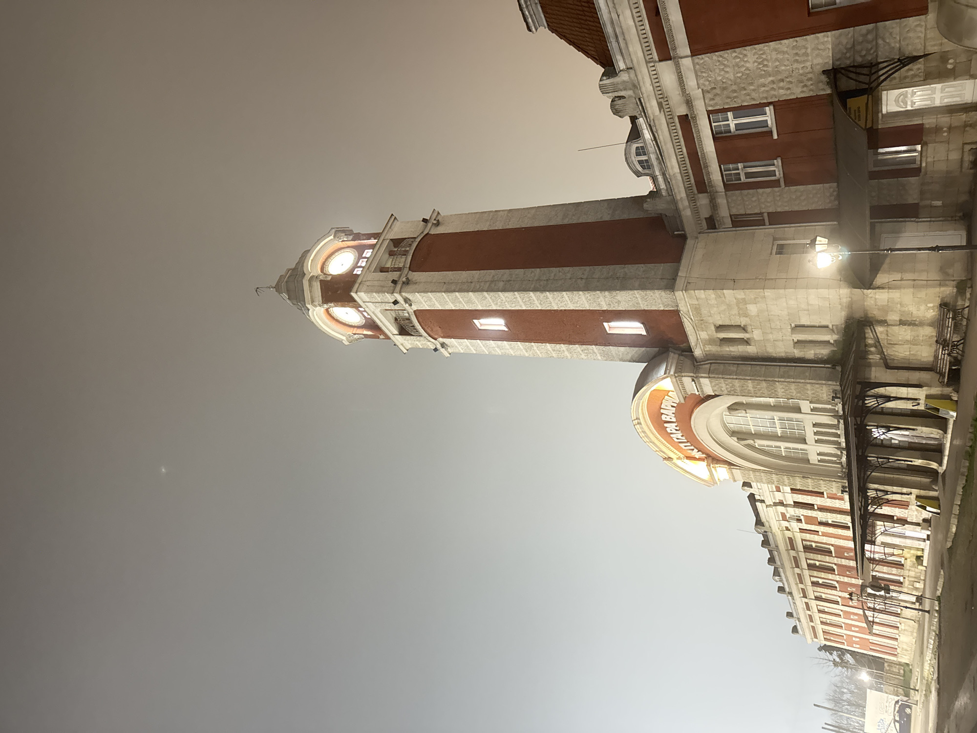 The image shows the Varna Clock Tower in Bulgaria at night. The tower is tall and slender with a red and white striped design. The top of the tower has illuminated clock faces. The surrounding building is a mix of red and white, and appears to be a train station. A street light is visible near the building. Bare trees are seen in the distance. The sky is a dark gray.