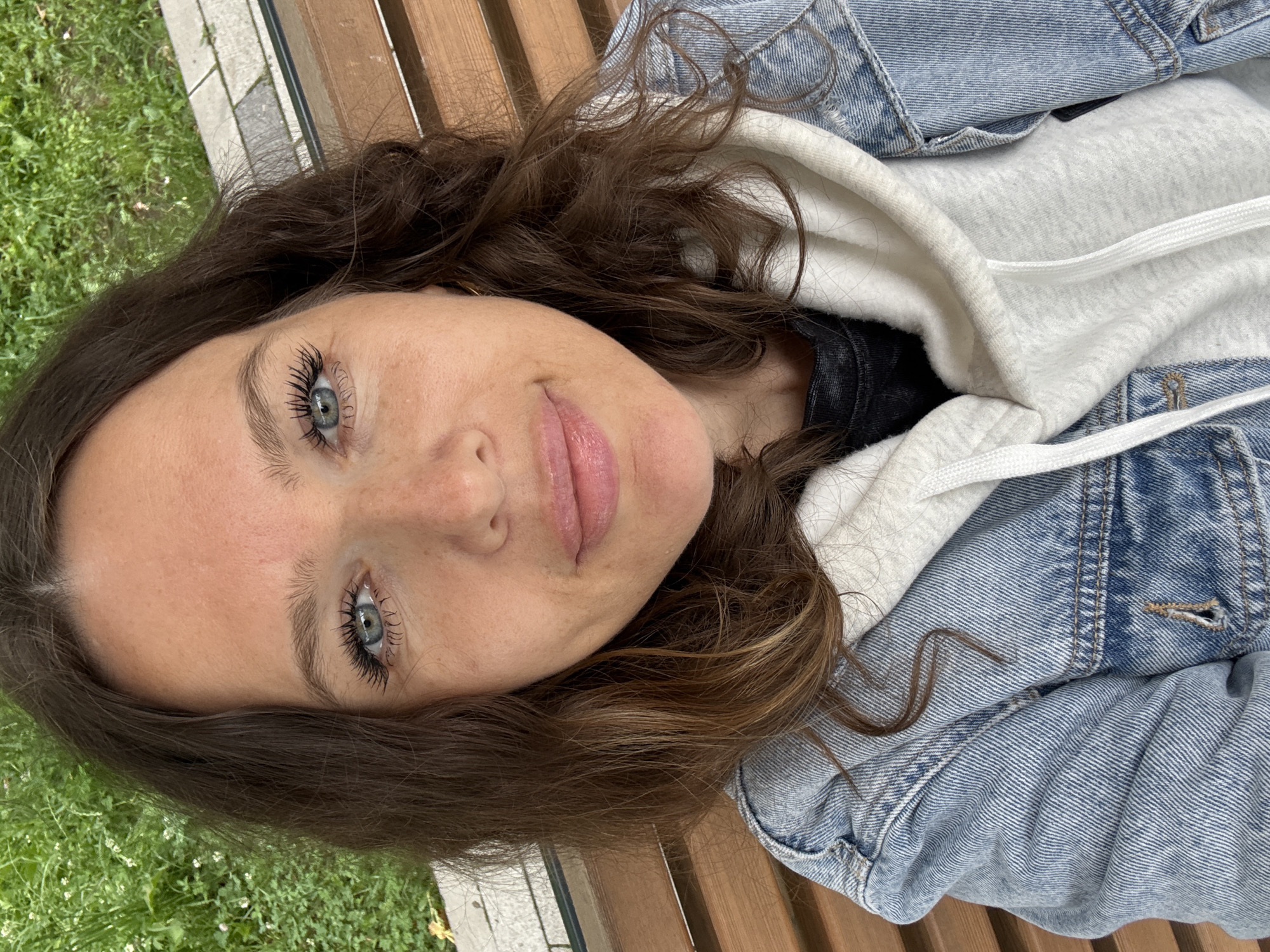 A close-up selfie of a woman with brown hair and blue eyes. She is wearing a denim jacket over a white hoodie. She is sitting on a wooden bench outdoors. The background consists of green grass and a paved area. The woman is looking directly at the camera and smiling slightly.