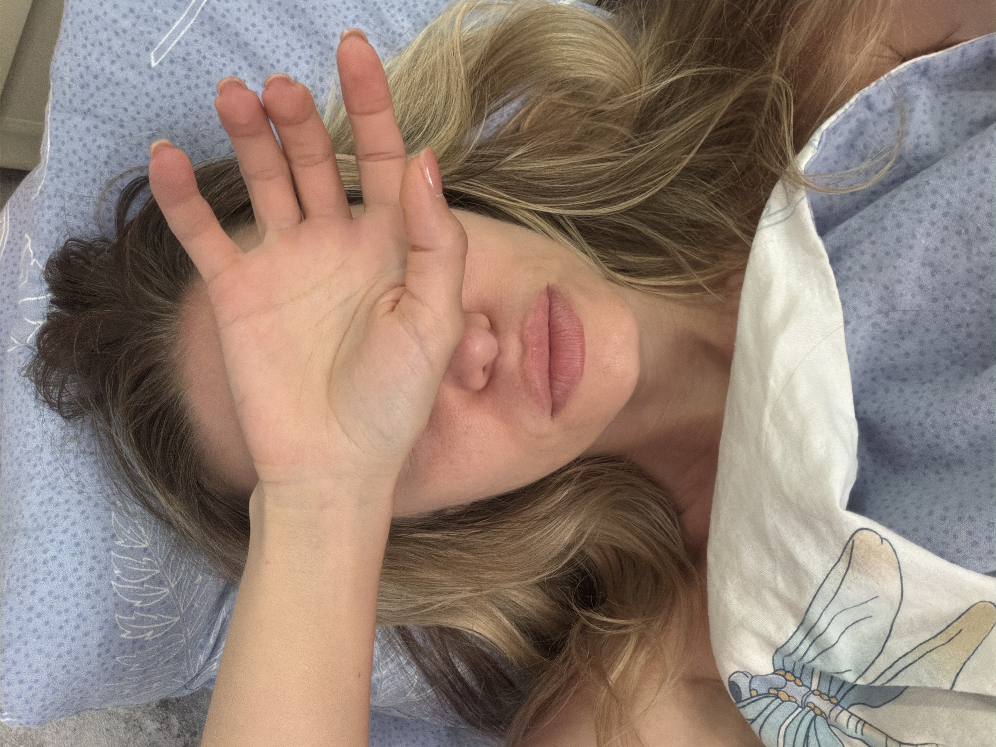 A woman's face is partially obscured by her hand as she lies in bed. Her hair is blonde and wavy, falling around her shoulders. She is wearing a white top with a blue and white patterned trim, tucked under a blue patterned duvet. The background consists of a blue patterned pillow. The image does not contain any elements that would indicate its location in the city of Sochi, Russia.
