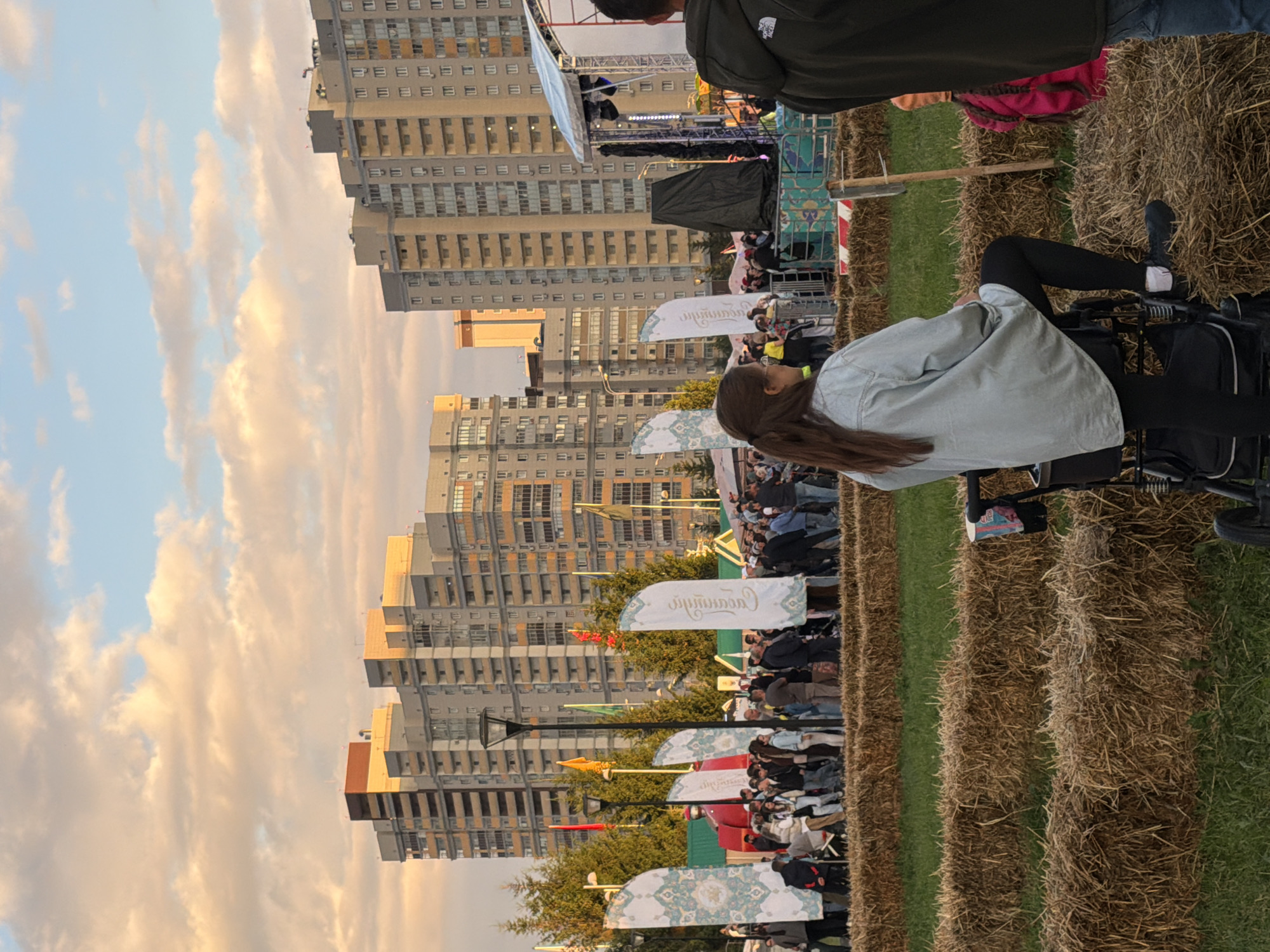 In an outdoor setting, a crowd gathers in front of tall, modern apartment buildings. The sky above is filled with wispy, pink-tinged clouds, suggesting either sunrise or sunset. In the foreground, rows of hay bales create a natural seating area. A woman, viewed from the back, is seated in a stroller, wearing a light blue denim jacket and black leggings. To her right, a child with braided hair and a bright pink backpack is partially visible.
Further back, a stage with lighting equipment is set up, hinting at a performance or event. Several large, decorative banners are displayed, featuring intricate patterns and text in what appears to be Arabic script. Among the crowd, people are dressed casually, enjoying the atmosphere. Trees dot the landscape, interspersed with lampposts and colorful flags. The overall scene depicts a community gathering in an urban environment, possibly a festival or an outdoor concert, under a dramatic sky.