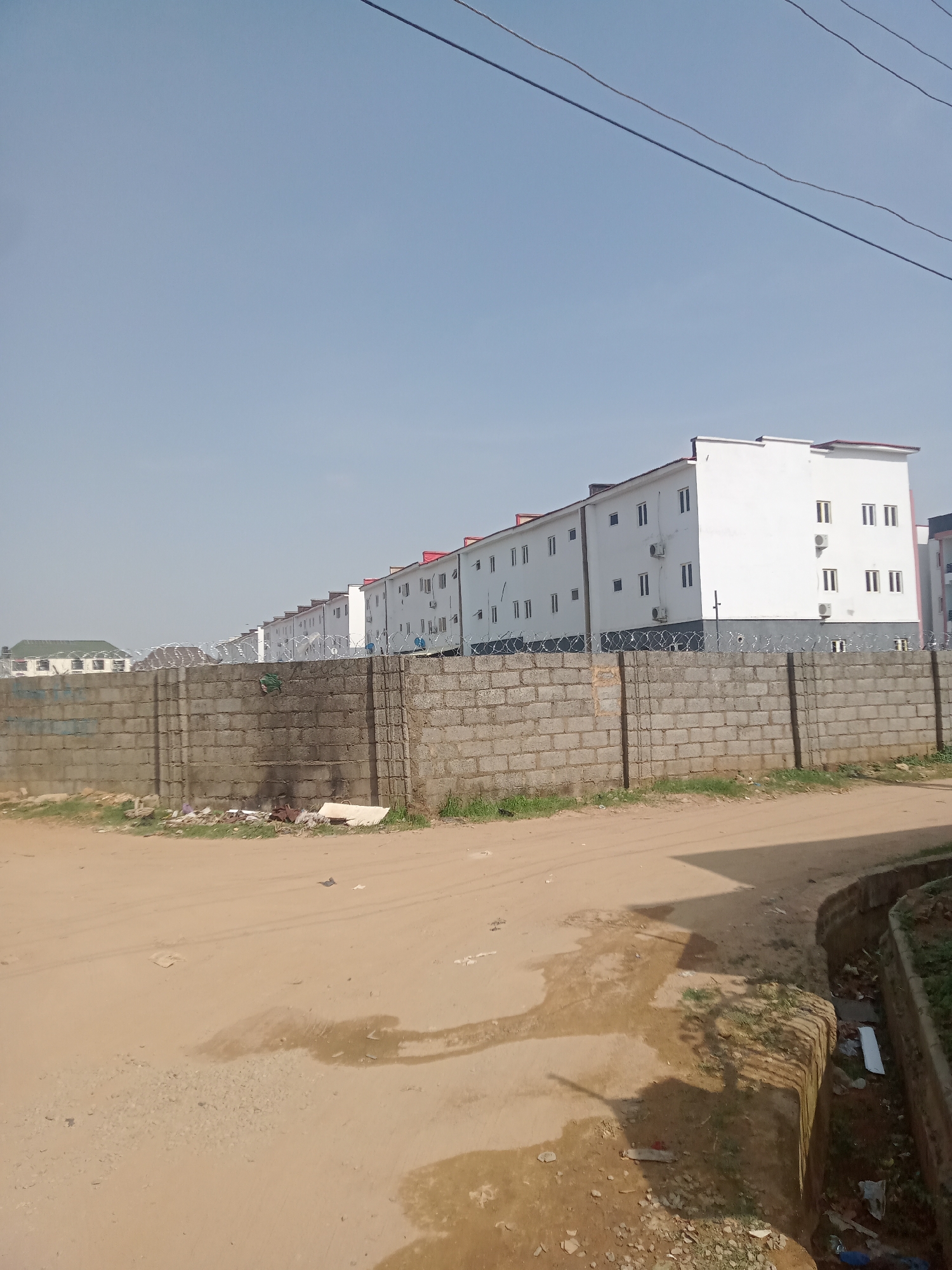 The image captures an outdoor scene in Gwarinpa, Nigeria, featuring a row of modern, white multi-story residential buildings under a clear, bright sky. These buildings, stretching into the distance from the midground, have red or dark grey sloped roofs, numerous dark windows, and visible exterior air conditioning units.
A tall, grey concrete block wall, topped with coiled razor wire, dominates the foreground, running horizontally across the frame. In front of this wall, an unpaved dirt road or path occupies the immediate foreground, showing patches of dry earth, sparse vegetation, and scattered litter. To the right of the road, a narrow drainage channel containing debris and some greenery is visible. In the far distance on the left, other structures with green roofs can be partially seen behind the main wall. Overhead, against the pale blue sky, thin power lines are discernible.
The scene depicts a quiet, static environment during the daytime, indicated by the bright, clear sky and strong illumination. No people are present, and no specific activities are occurring. The overall impression is one of a developing suburban area or a newly constructed housing estate, characterized by functional architecture and basic infrastructure, consistent with the Gwarinpa context. There is no visible text in the image.