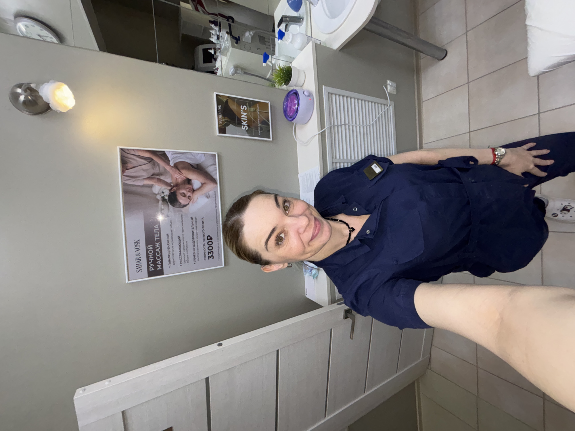 A smiling woman, likely a professional in a salon or spa setting, takes a selfie indoors. She has brown hair pulled back and wears a dark blue uniform or scrubs, along with a watch on her left wrist.
The background features light grey walls and a beige-ttiled floor. On the wall to the left, a sconce emits a warm light. A white door or cabinet with vertical panels is partially visible in the foreground. In the mid-ground, there's a counter area with a white sink, faucet, and various items like bottles, a small potted plant, and a purple wax warmer. A large mirror above the sink reflects parts of the room, including some equipment and a wall clock. A ventilation grate is visible below the counter.
Two framed posters are displayed on the wall. The larger poster on the left, branded "SAHAR & VOSK," advertises "РУЧНОЙ МАССАЖ ТЕЛА" (Hand Body Massage), listing types such as "ЛИМФОДРЕНАЖНЫЙ" (lymphatic drainage), "РАССЛАБЛЯЮЩИЙ" (relaxing), and "ЛЕЧЕБНО-ОЗДОРОВИТЕЛЬНЫЙ" (therapeutic-healing). It states the "СТОИМОСТЬ ПЕРВОГО ВИЗИТА" (Cost of first visit) as "3300P" and includes an image of a woman receiving a massage. The smaller poster to the right also bears the "SAHAR & VOSK" brand and the word "SKIN'S."
The scene conveys a clean, professional, and welcoming environment, likely a beauty or wellness establishment. The lighting is artificial, suggesting an indoor setting at an indeterminate time of day.