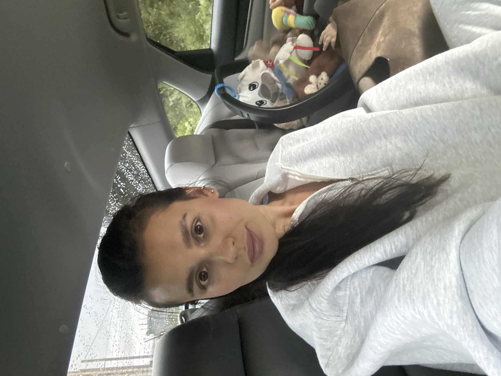 A woman is sitting in the driver's seat of a car, looking at the camera. Her hair is pulled back and she is wearing a light gray hooded sweatshirt. In the back seat, a baby is visible in a car seat with several toys. The weather outside appears to be rainy, with water droplets visible on the car's windows. The setting is likely a residential area or a street in Perm, Russia, given the cloudy and wet conditions.