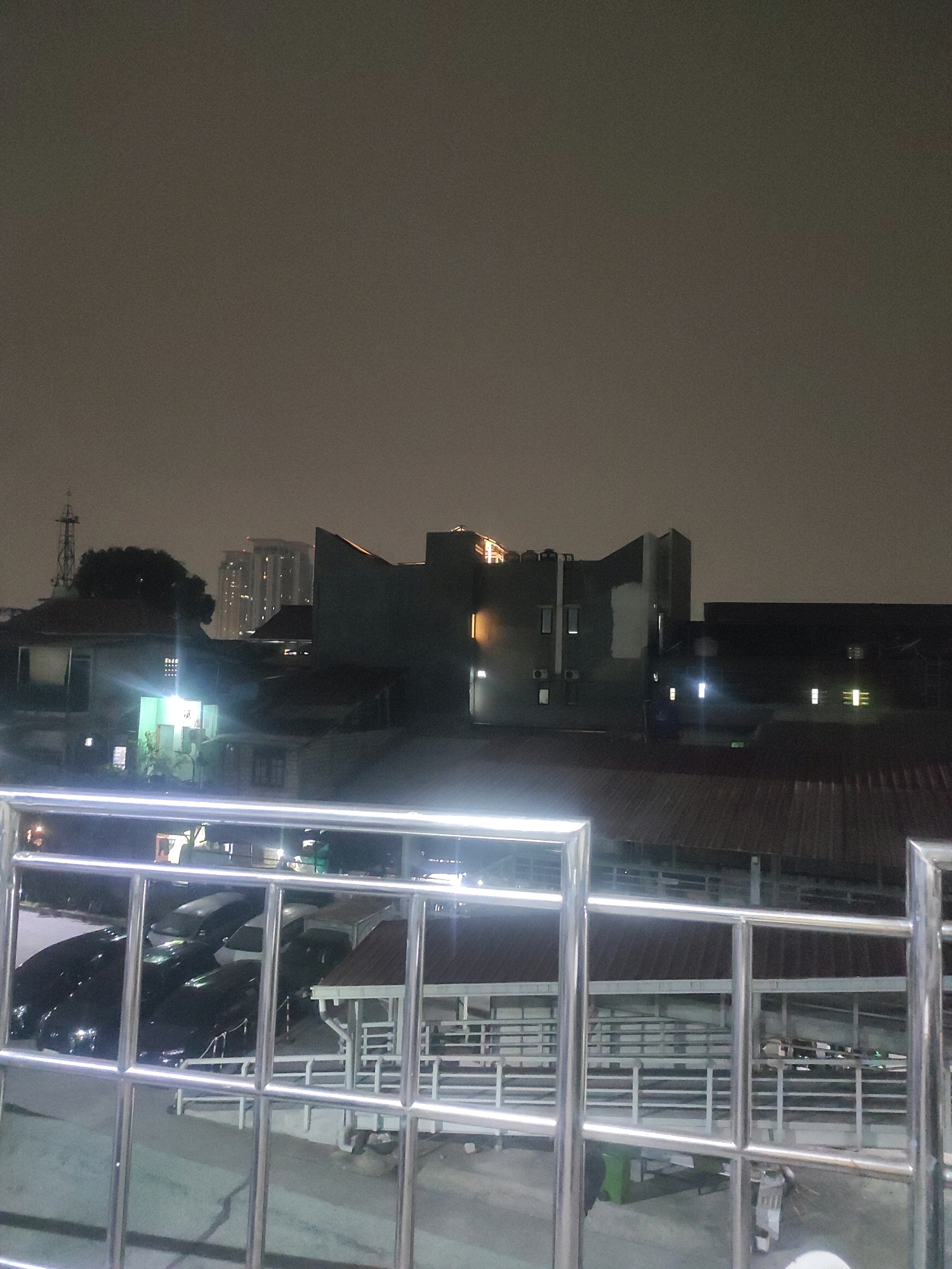 The image captures a night scene in South Jakarta, Indonesia, showing a collection of buildings and a parking area viewed through a metal railing. The sky is overcast and dark, with some distant lights from taller buildings visible.
In the foreground, a silver metal railing with square openings dominates the lower portion of the image, suggesting a vantage point from a balcony or elevated walkway.
Beyond the railing, a parking area is visible. Several cars, primarily dark-colored, are parked in rows. Some have their headlights on, casting beams onto the pavement. A white car is parked amidst the darker vehicles. Structures with covered roofs, possibly carports or small buildings, are interspersed within the parking area.
Further in the background, a cluster of buildings of varying heights and architectural styles are present. One building, dark gray in color, features a distinct angular design and several illuminated windows. To its left, a smaller building with a brightly lit window casts a vibrant green light outwards. A telecommunications tower stands tall in the far left background. In the distance, a modern high-rise building with numerous illuminated windows stands out against the dark sky.
The overall impression is of a dense urban environment at night, characterized by artificial lighting illuminating various structures and vehicles. The image is grainy, consistent with low-light photography.