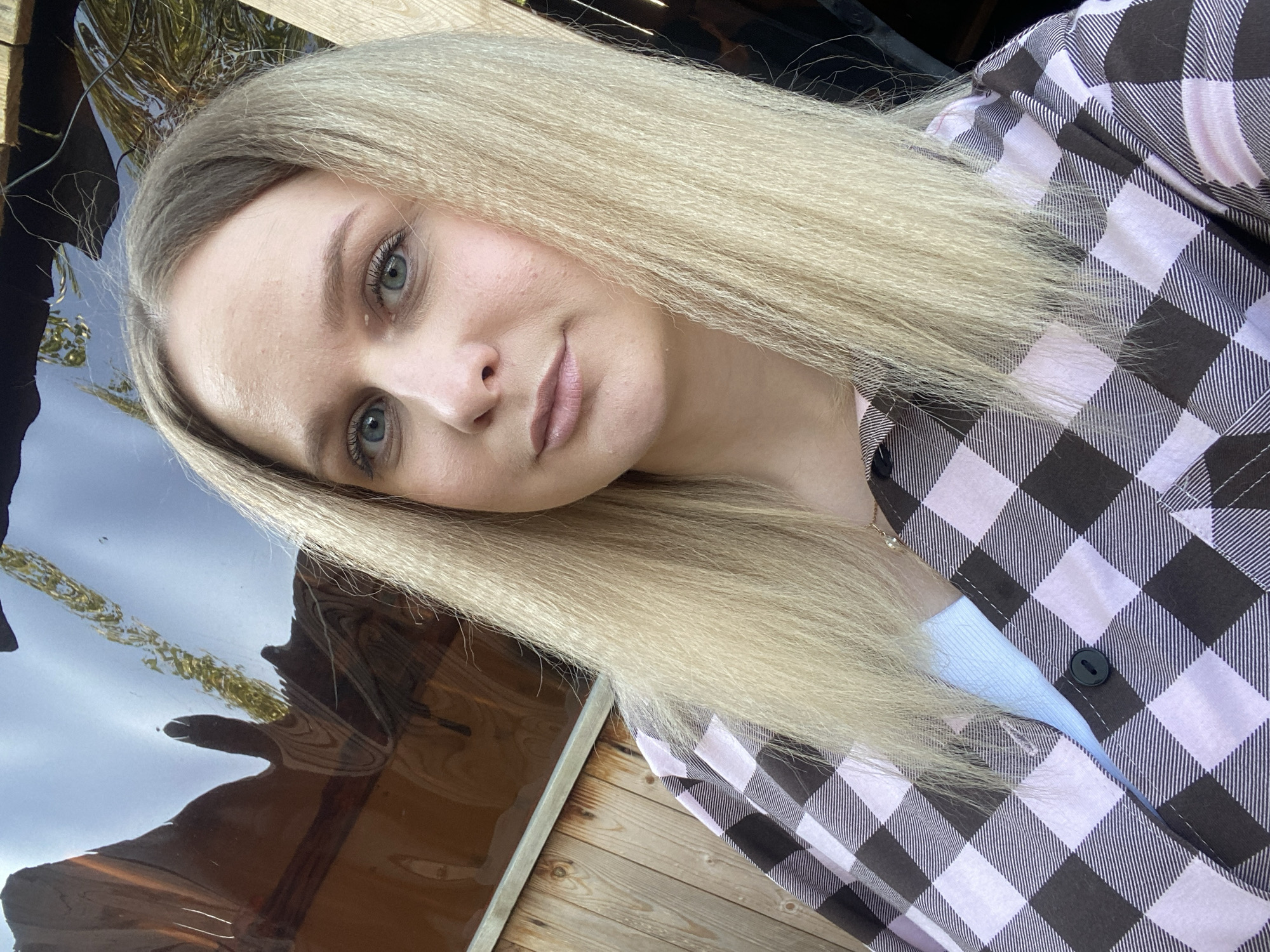 A close-up selfie features a young woman with long, textured blonde hair and light blue-grey eyes, looking directly at the camera with a neutral to slightly contemplative expression. Her hair appears crimped or finely waved. She has fair skin, with visible natural skin texture, and is wearing light makeup, including a light pink lipstick.
She is dressed in a black and light pink (or lavender) plaid button-up shirt, worn open over a light blue or white undershirt. A dark button is visible on her plaid shirt.
The setting is outdoors, with the image captured at an angle. The background reveals reflections that suggest a body of water, possibly a lake or river, reflecting a bright sky and some distant structures or foliage. Wooden planks or decking are visible in the lower and left portions of the frame, indicating a structure near the water. The overall impression is a sunny daytime environment. There is no visible text in the image. The location is Kauchuk Stantsiya, Russia.